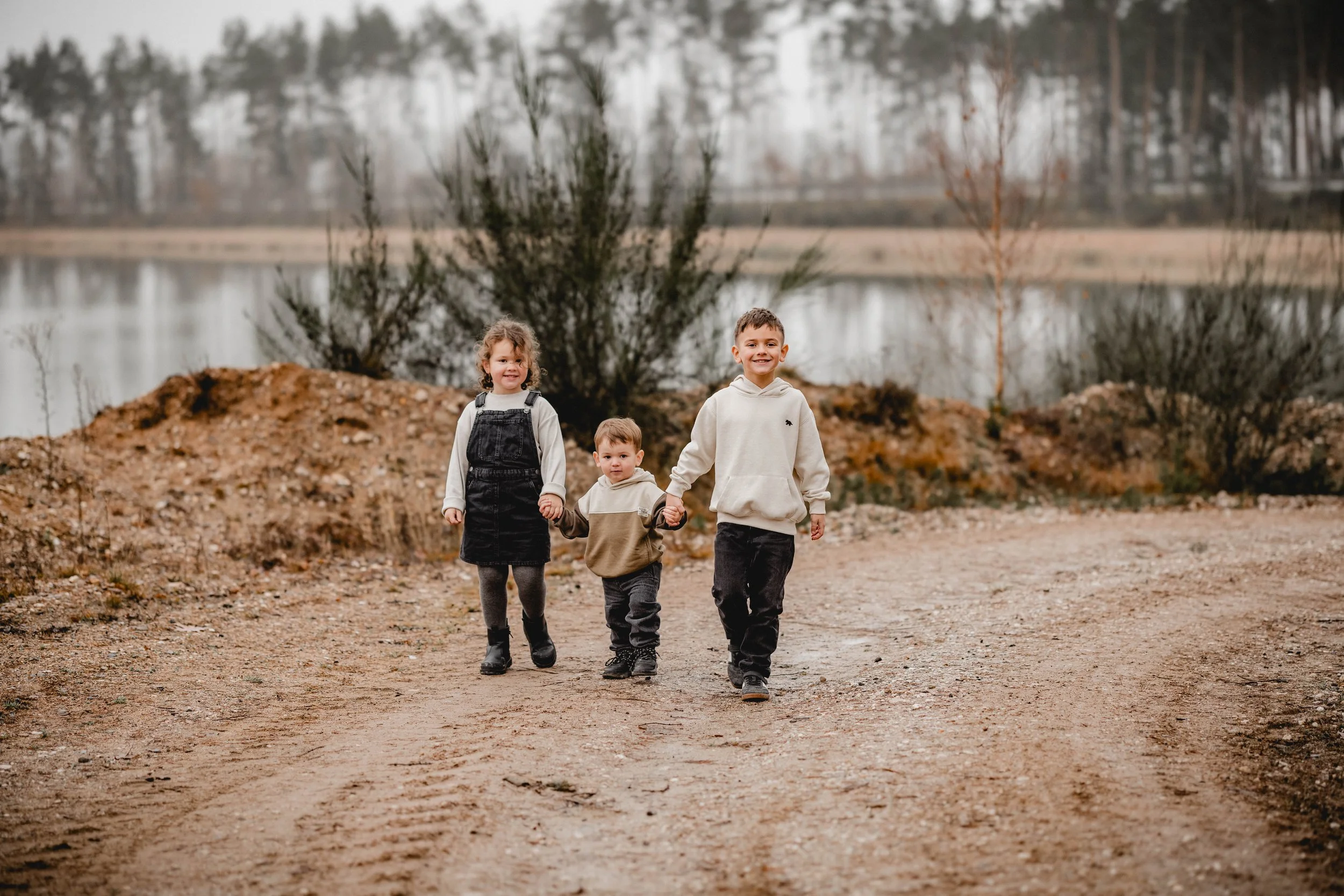 Drei lachende Kinder gehen Hand in Hand auf einem Weg entlang eines Sees im Herbst.