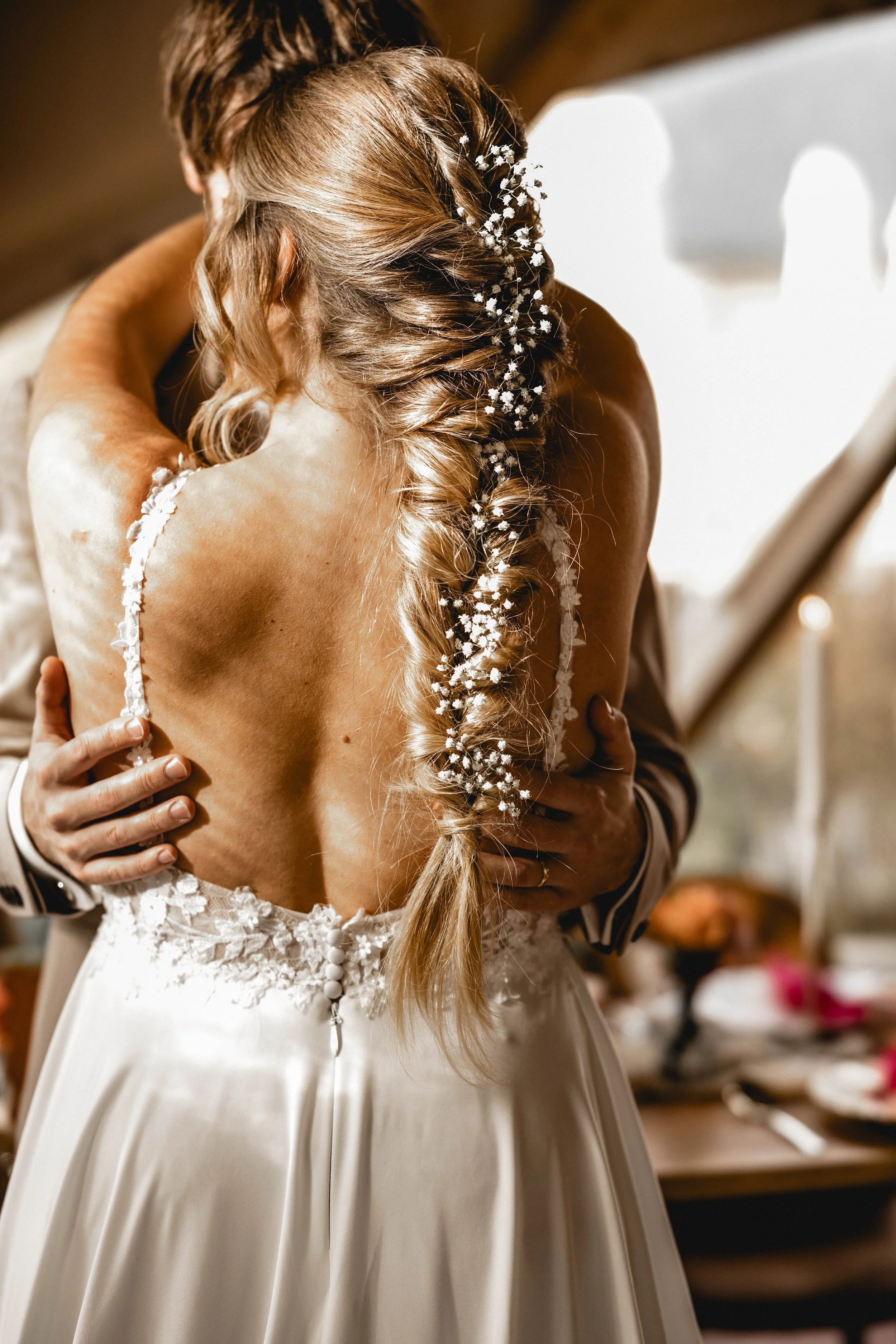 Braut mit einem voluminösen Kleid und einer geflochtenen Hochsteckfrisur mit kleinen weißen Blumen. Ein Mann umarmt sie.
