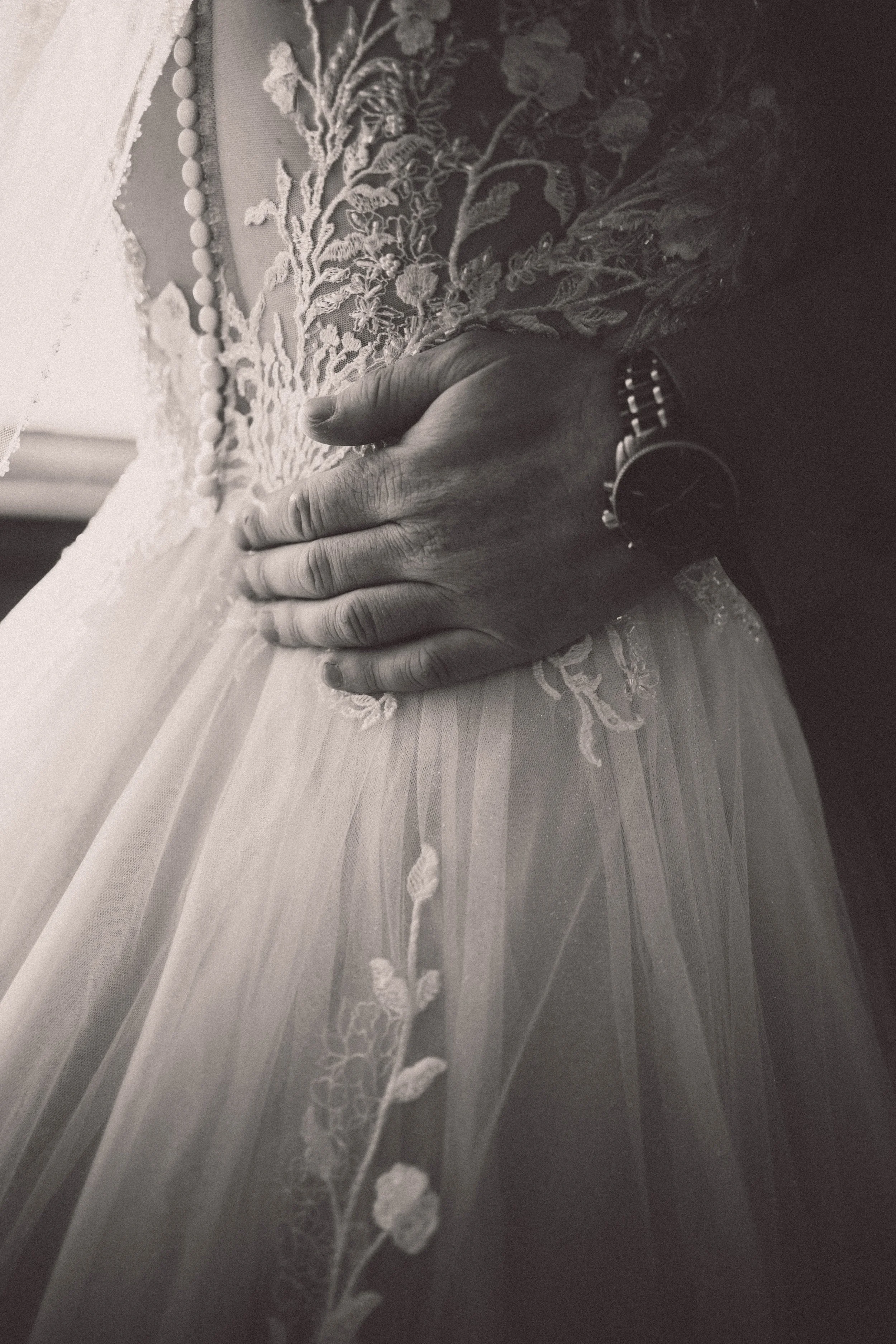 Nahaufnahme einer Hand, die auf das lace-Design eines Brautkleides liegt, mit einer Uhr am Handgelenk, schwarz-weiß.