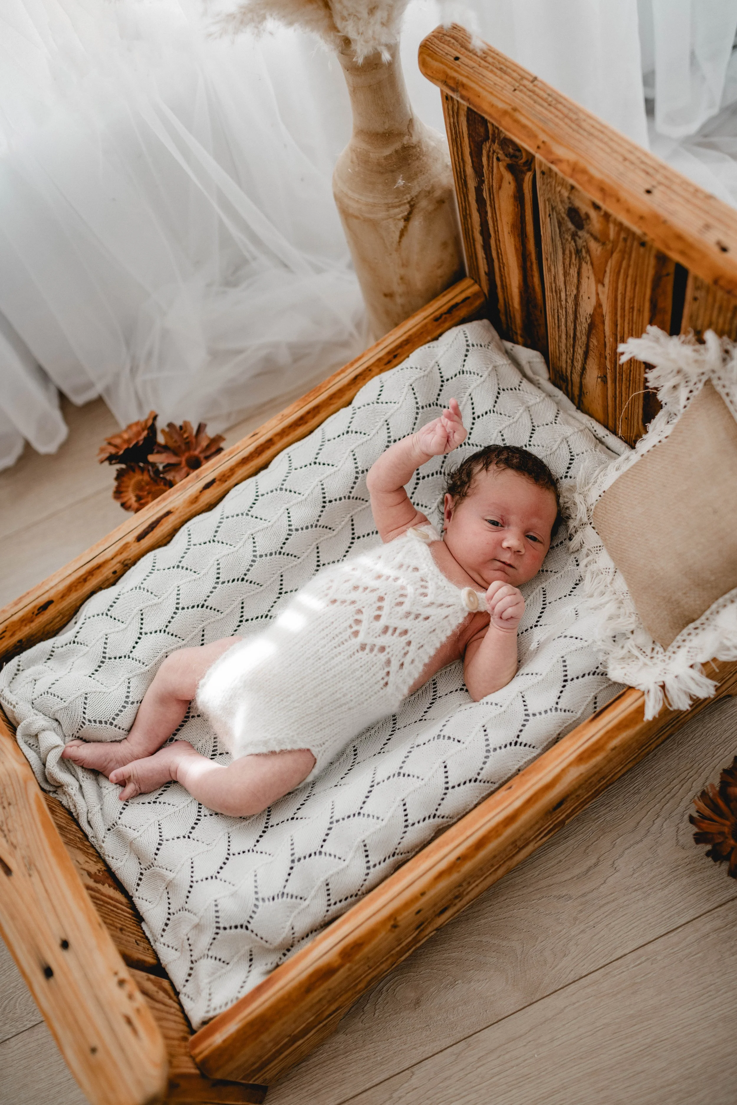 Ein Baby liegt in einem Holz-Etagenbett, trägt ein weißes gestricktes Kleid und schaut in die Kamera. Im Hintergrund sind weiße Vorhänge und ein Keramikvase.
