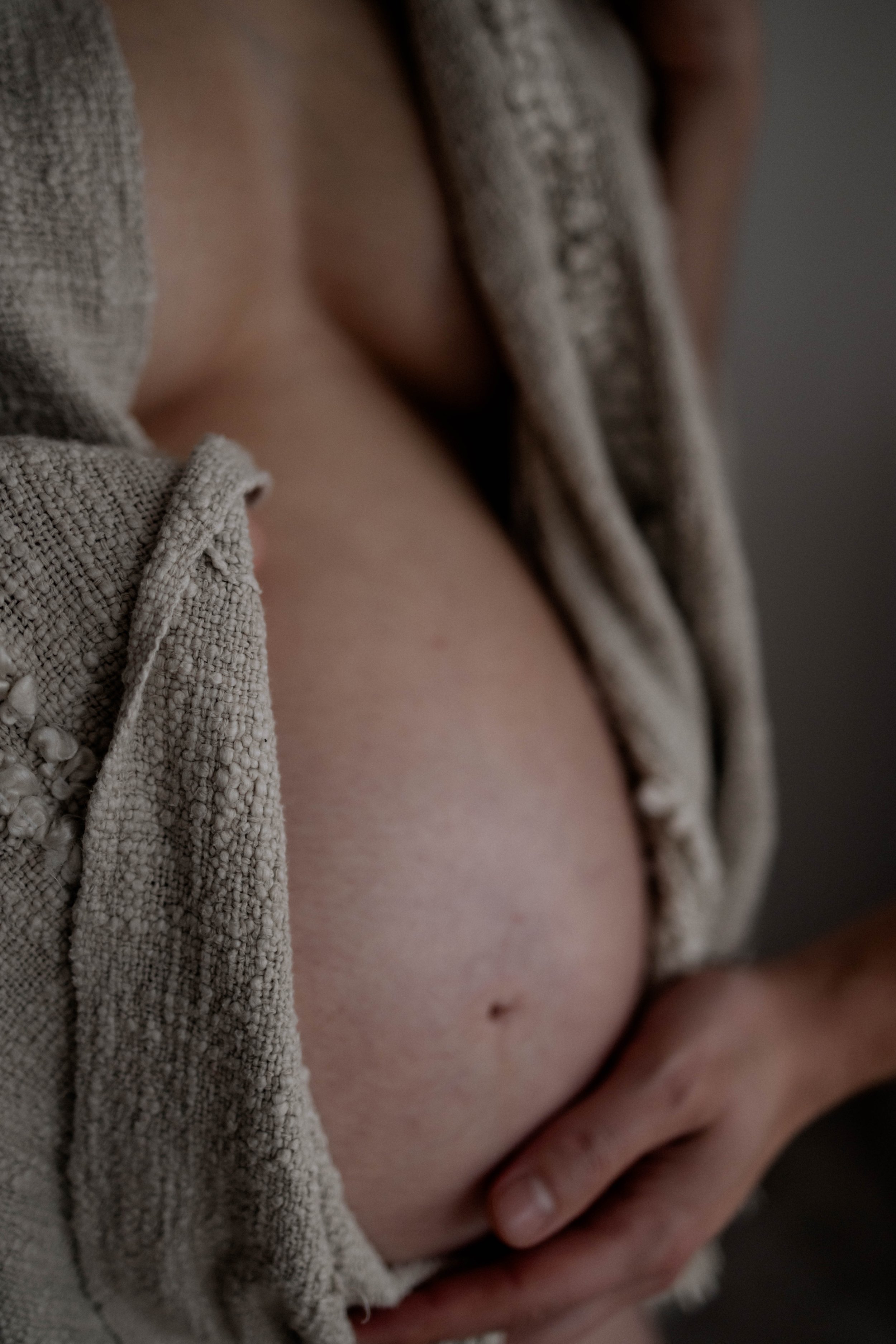 Nahaufnahme eines Bauches mit einer Hand, die ihn stützt, teilweise bedeckt von einem grauen, strukturierten Stoff.