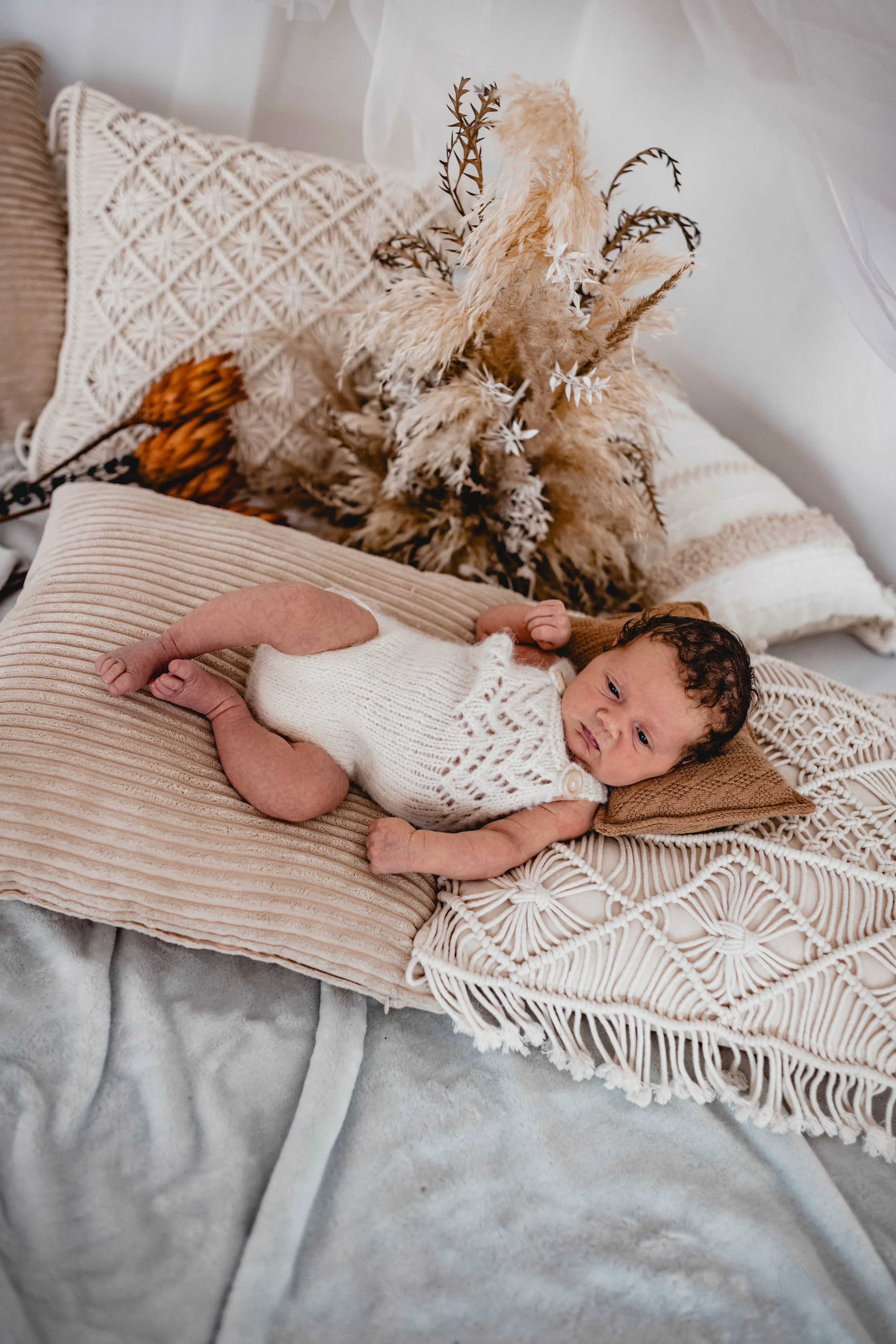 Ein neugeborenes Baby liegt auf einem beige Kissen und hat eine weiße Strickweste an, umgeben von dekorativen Kissen und einem Trockenblumenstrauß im Hintergrund.