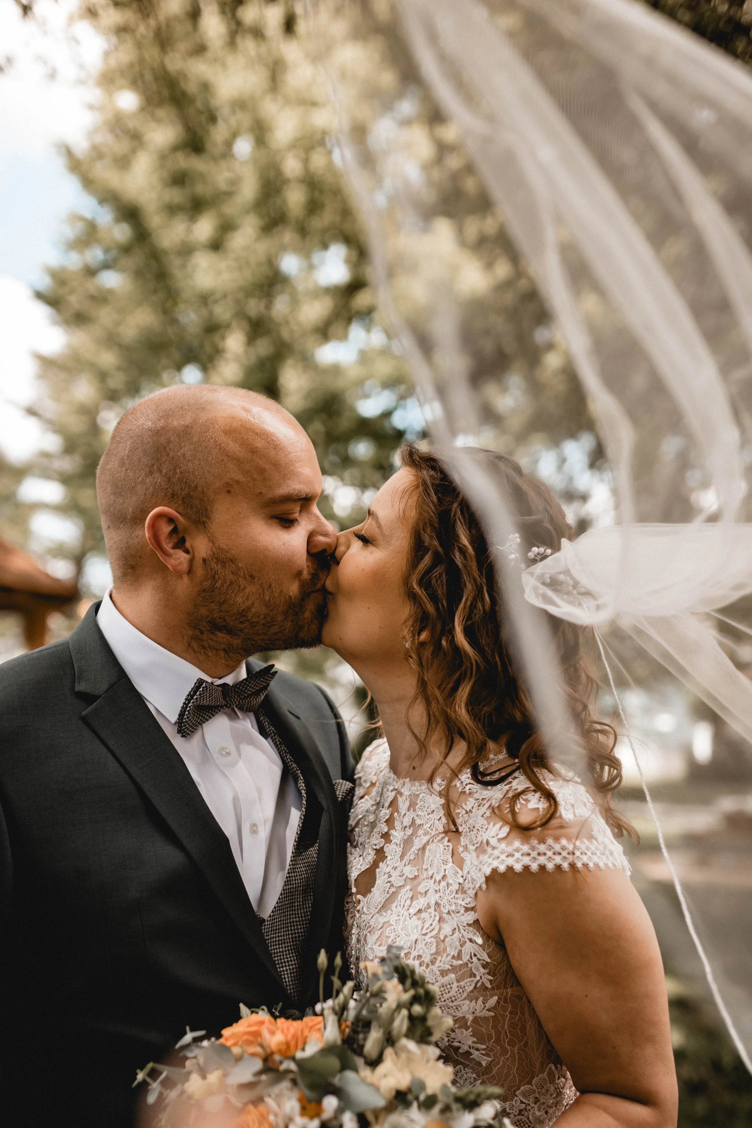 Ein Brautpaar küsst sich bei einer Hochzeit im Freien, die Braut trägt ein Spitzenkleid mit Schleier, der Bräutigam trägt einen schwarzen Anzug mit Fliege, im Hintergrund Bäume und Himmel.