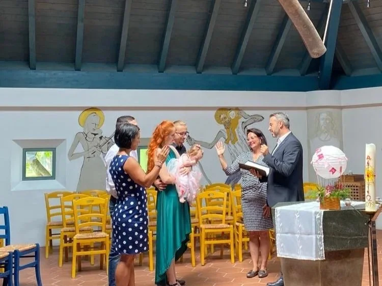 Eine Gruppe von Menschen beteiligt sich an einer Taufe in einem Kirchenraum, während ein Priester ein Buch liest und die Teilnehmer mit Handgesten segnet.