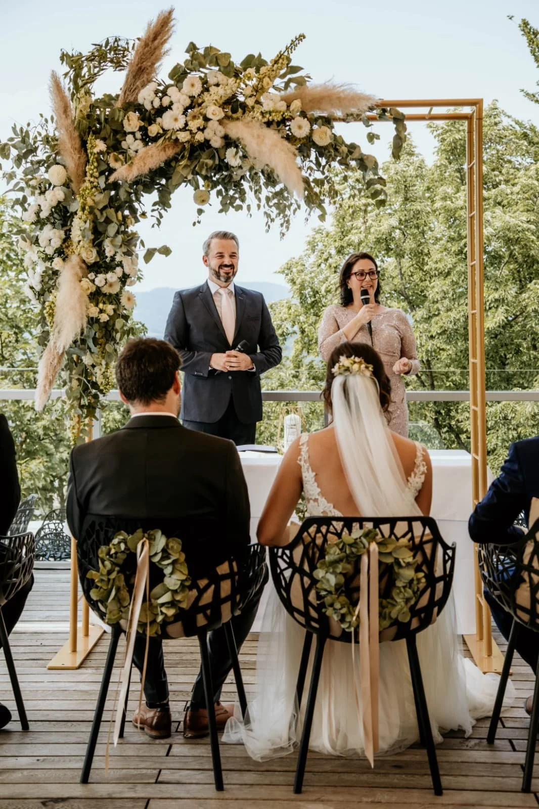 Ein Hochzeitszeremonie im Freien auf einer Holzterrasse mit einer goldenen Blumenrahmen und weißen Blumen. Das Brautpaar sitzt, die Braut trägt ein weißes Kleid und ein Blumenhaarband. Der Bräutigam trägt einen schwarzen Anzug. Zwei Personen, eine Frau hält eine Mikrofon, sprechen, während die Gäste zuhören.