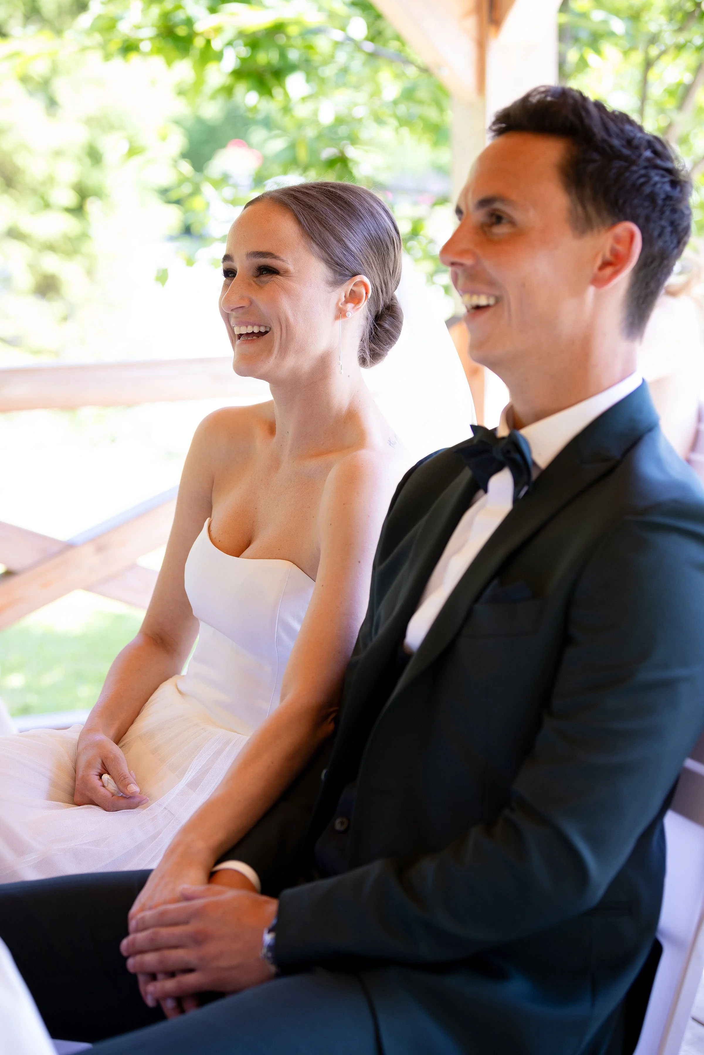 Ein lachendes Brautpaar bei einer Hochzeit, sitzend nebeneinander, im Freien mit grünen Bäumen im Hintergrund.