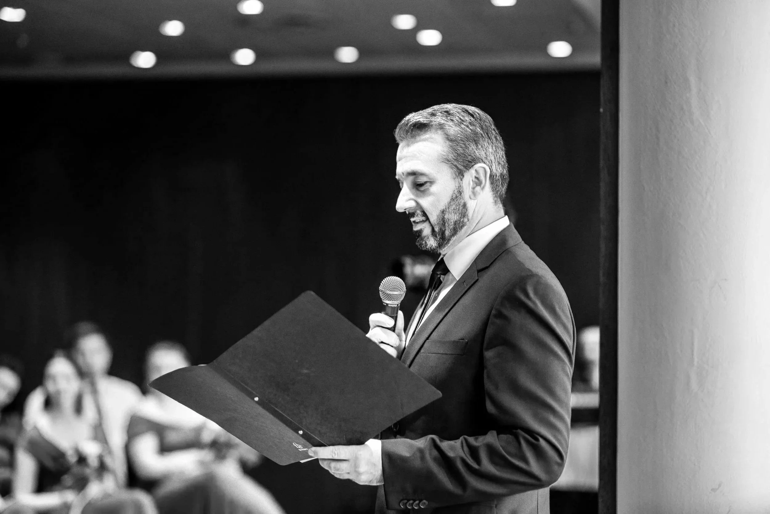 Ein Mann in einem Anzug liest aus einem Ordner vor, während er eine Mikrofon hält. Im Hintergrund sitzen Menschen und hören zu.