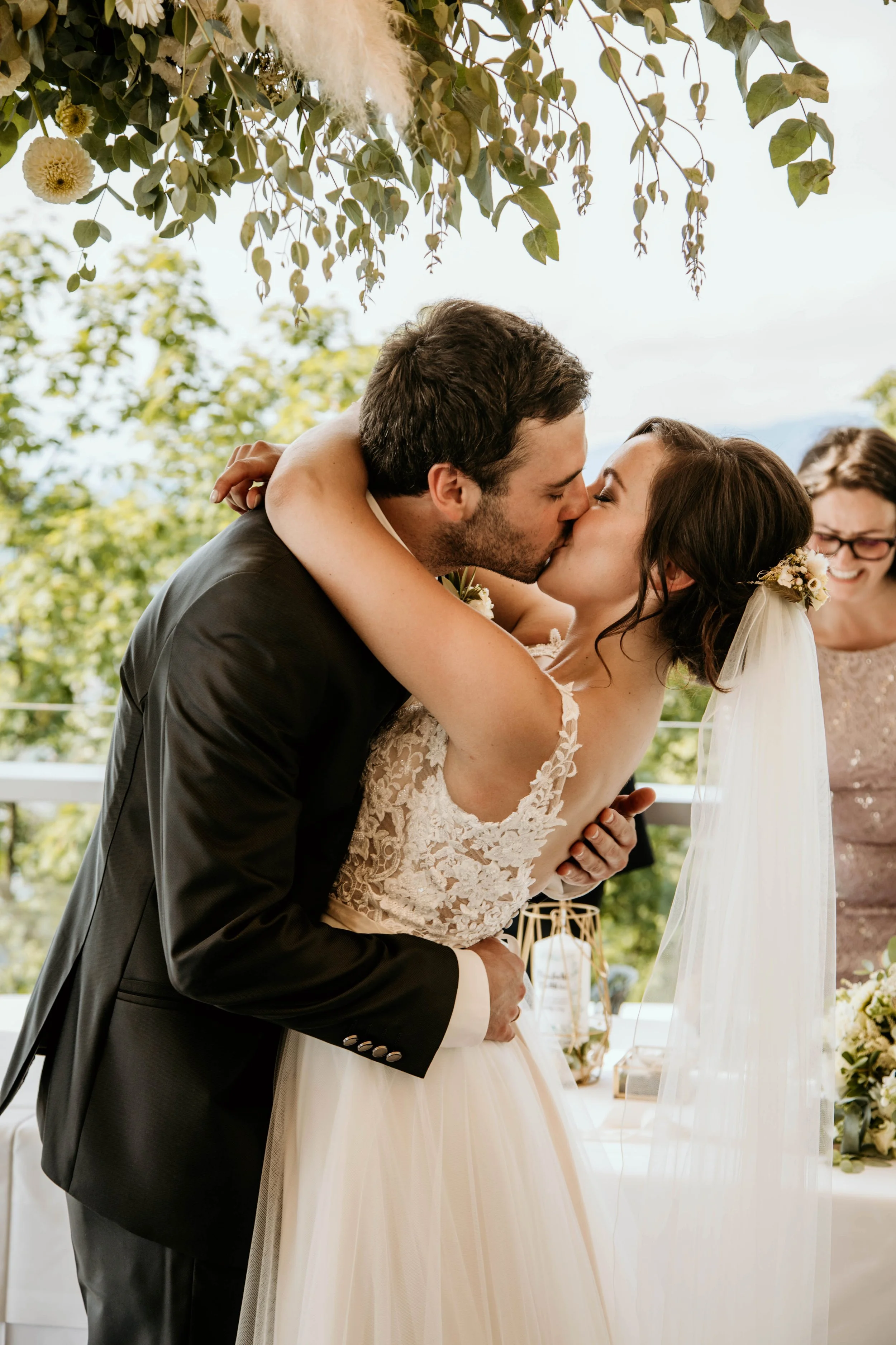 Ein Hochzeitskuss zwischen einem Bräutigam in schwarzem Anzug und einer Braut in einem weißen Kleid mit Spitze, mit einer lachenden Frau im Hintergrund bei einer Hochzeit im Freien.