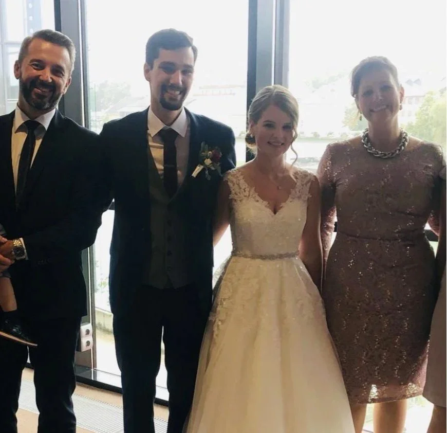 Gruppe von fünf Menschen, bei einer Hochzeitsfeier, im Raum mit großen Fenstern, freundliche Atmosphäre, das Brautpaar in der Mitte, die anderen im eleganten Outfit.