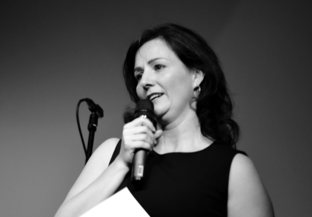 Eine Frau beim Sprechen auf der Bühne, hält ein Mikrofon in der Hand, im Hintergrund ein schwarzer Vorhang.