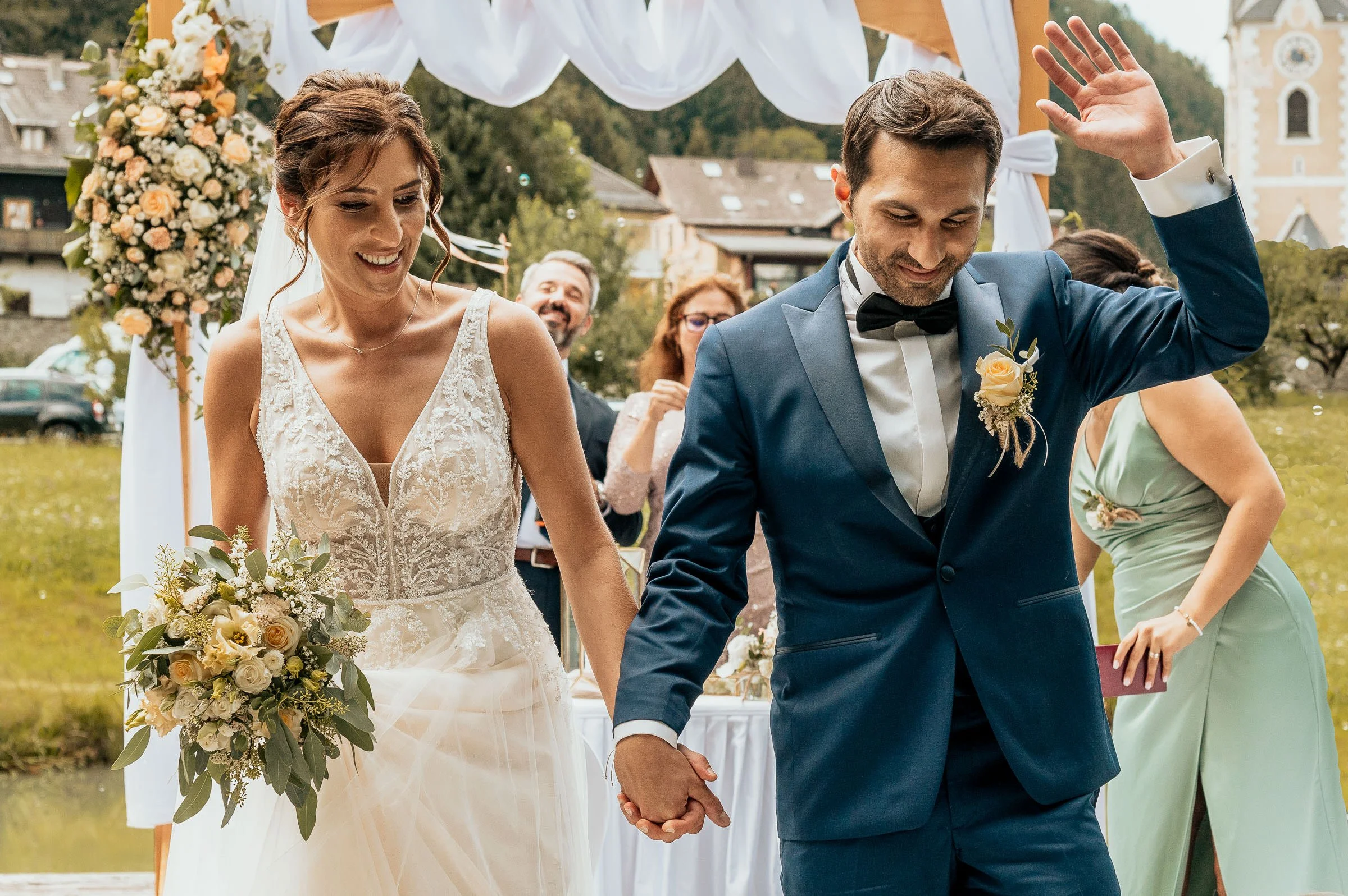 Ein Brautpaar hält während einer Hochzeit im Freien die Hände, die Braut trägt ein weißes Hochzeitskleid und hält einen Blumenstrauß, der Bräutigam trägt einen blauen Anzug und eine Fliege, im Hintergrund sind Gäste und eine Kirche sichtbar.