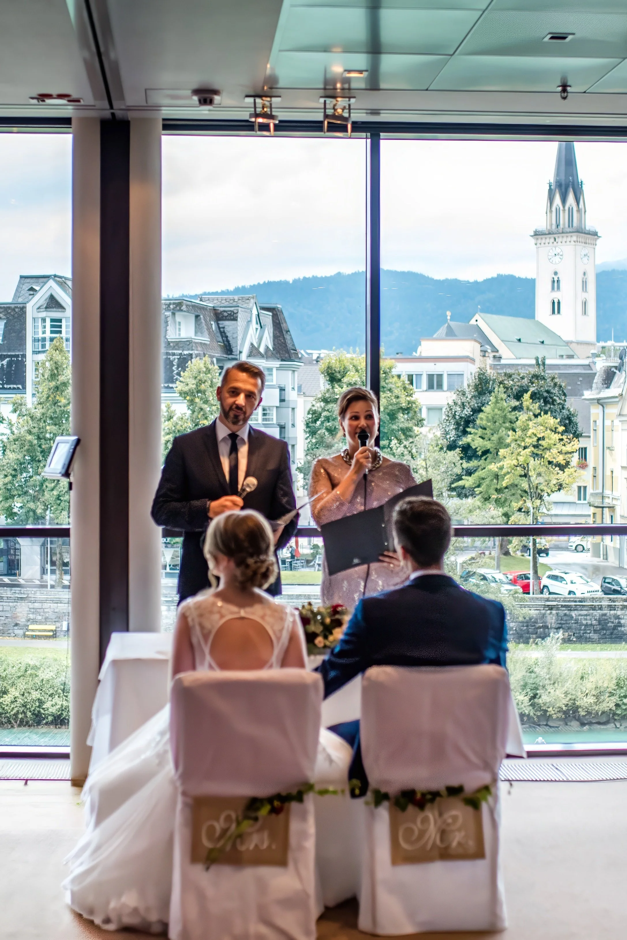 Hochzeitspaar sitzt an einem Tisch, im Hintergrund stehen ein Mann und eine Frau mit Mikrofonen bei einer Zeremonie mit Blick auf eine Stadt mit Kirchen und Gebäuden, vor einer großen Fensterfront.