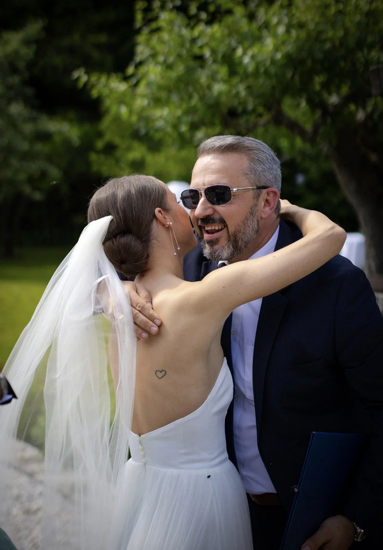Braut und ein Mann bei einer Umarmung auf einer Hochzeit im Freien, lachend, mit Sonnenbrille, im Hintergrund Bäume.