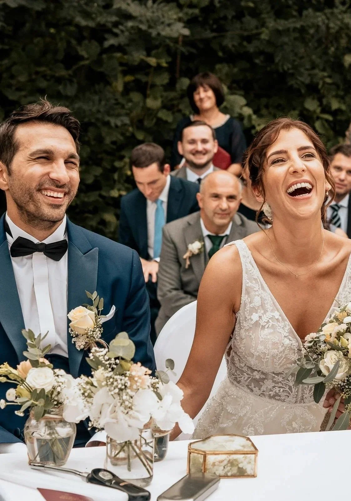 Ein glückliches Brautpaar bei ihrer Hochzeit, lachend, mit Freunden im Hintergrund in einem Garten