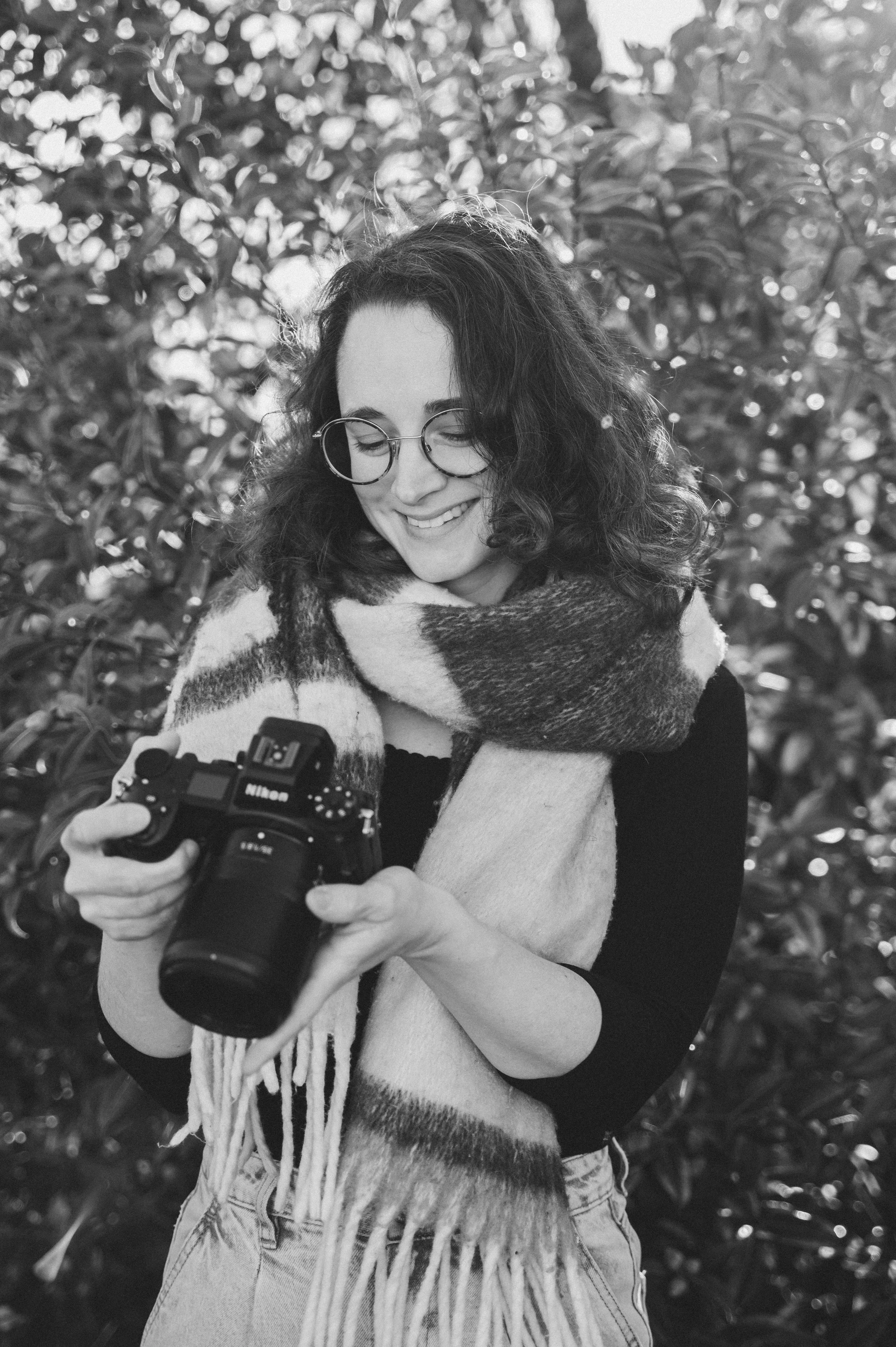 Une femme souriante avec des lunettes rondes, portant une écharpe à carreaux, regarde un appareil photo Nikon qu'elle tient dans ses mains, en extérieur avec un fond de feuillages.