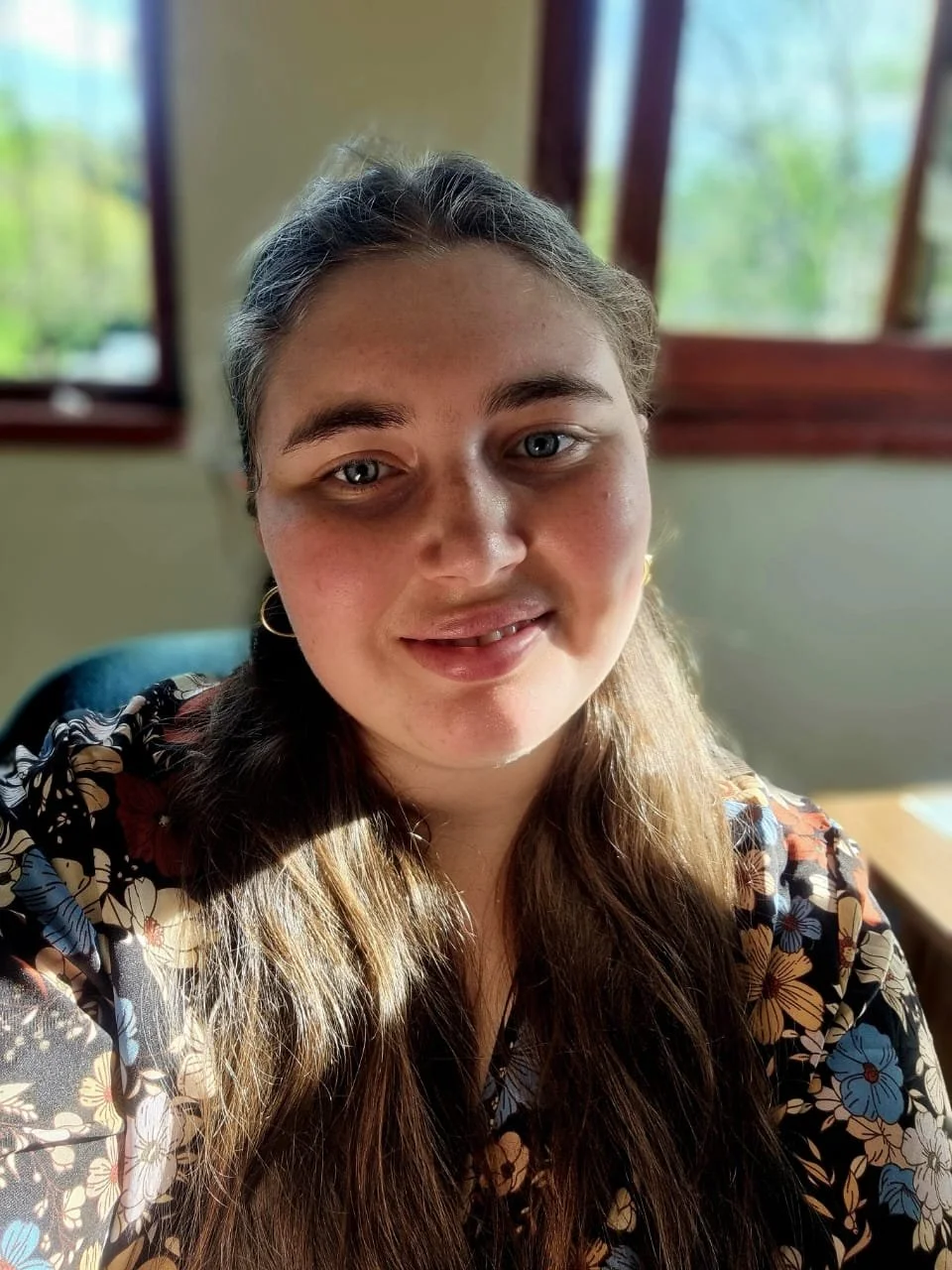 Close-up of a young woman with long light brown hair, wearing a floral patterned top and gold hoop earrings, smiling in a room with windows showing greenery outside.