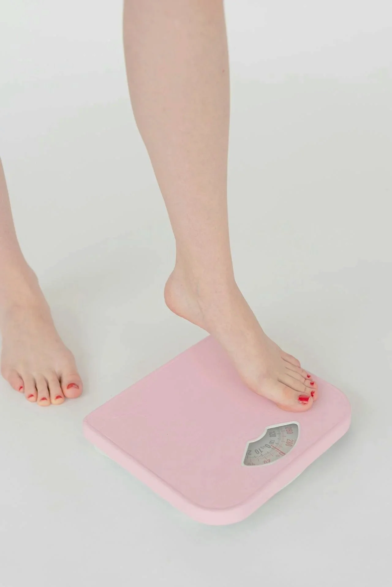 Person standing barefoot with one foot on a pink bathroom scale, with the other foot nearby on a white surface, in a bright setting.