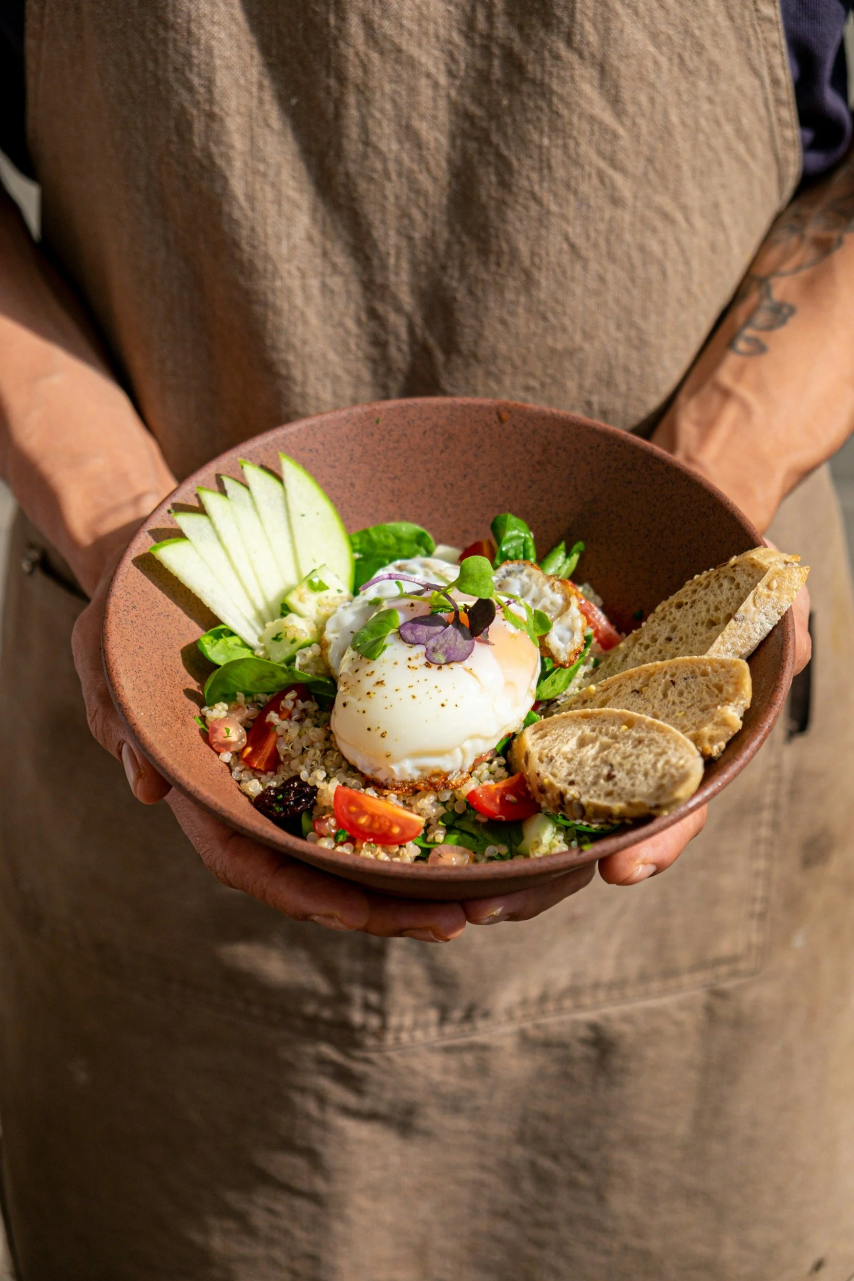 A person holding a bowl of quinoa salad with cherry tomatoes, cucumber slices, microgreens, an poached egg, and slices of baguette.