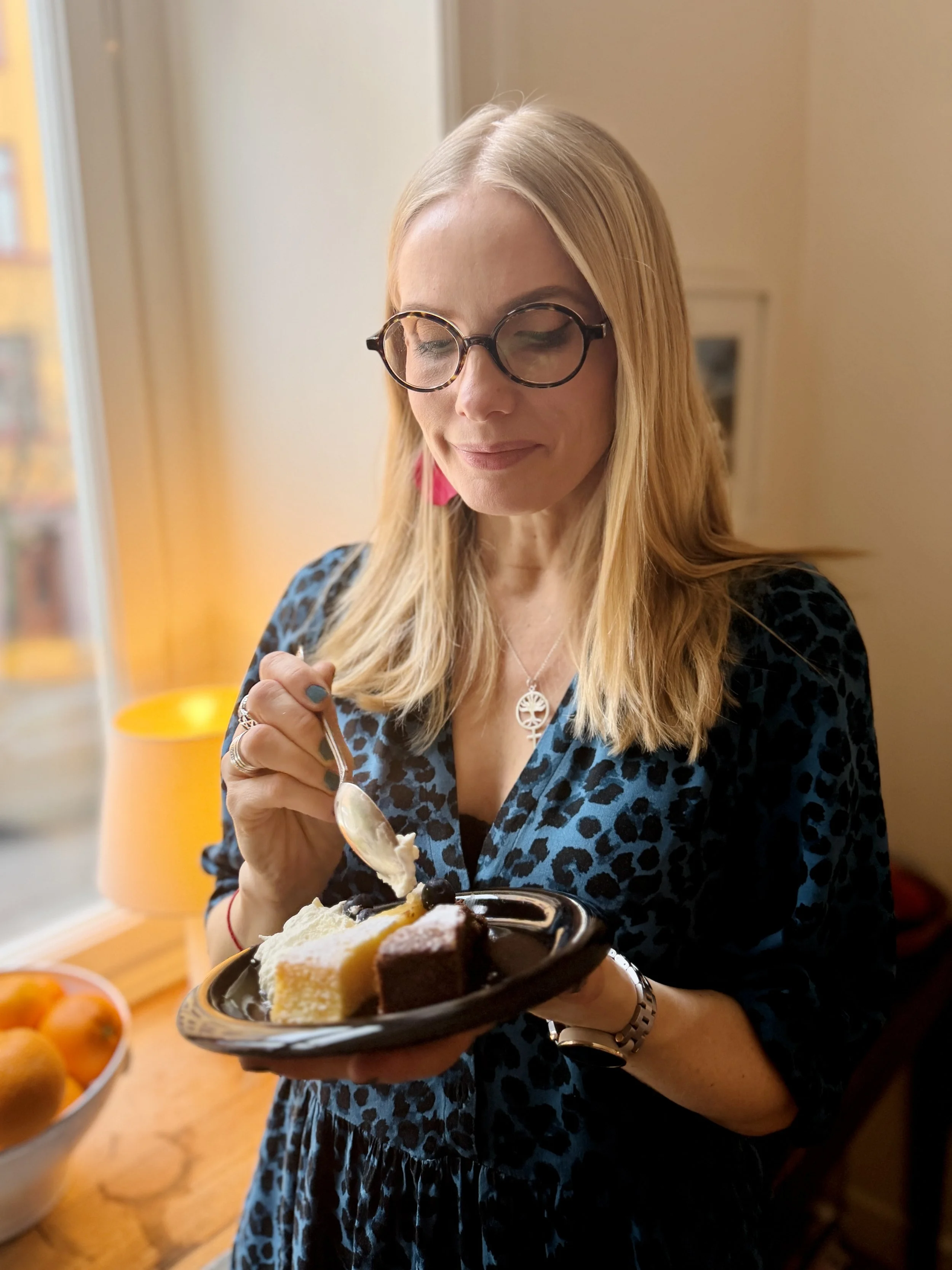 Woman with blonde hair, glasses, and a blue leopard print dress, holding a black plate with various slices of cake, in a cozy indoor setting near a window.