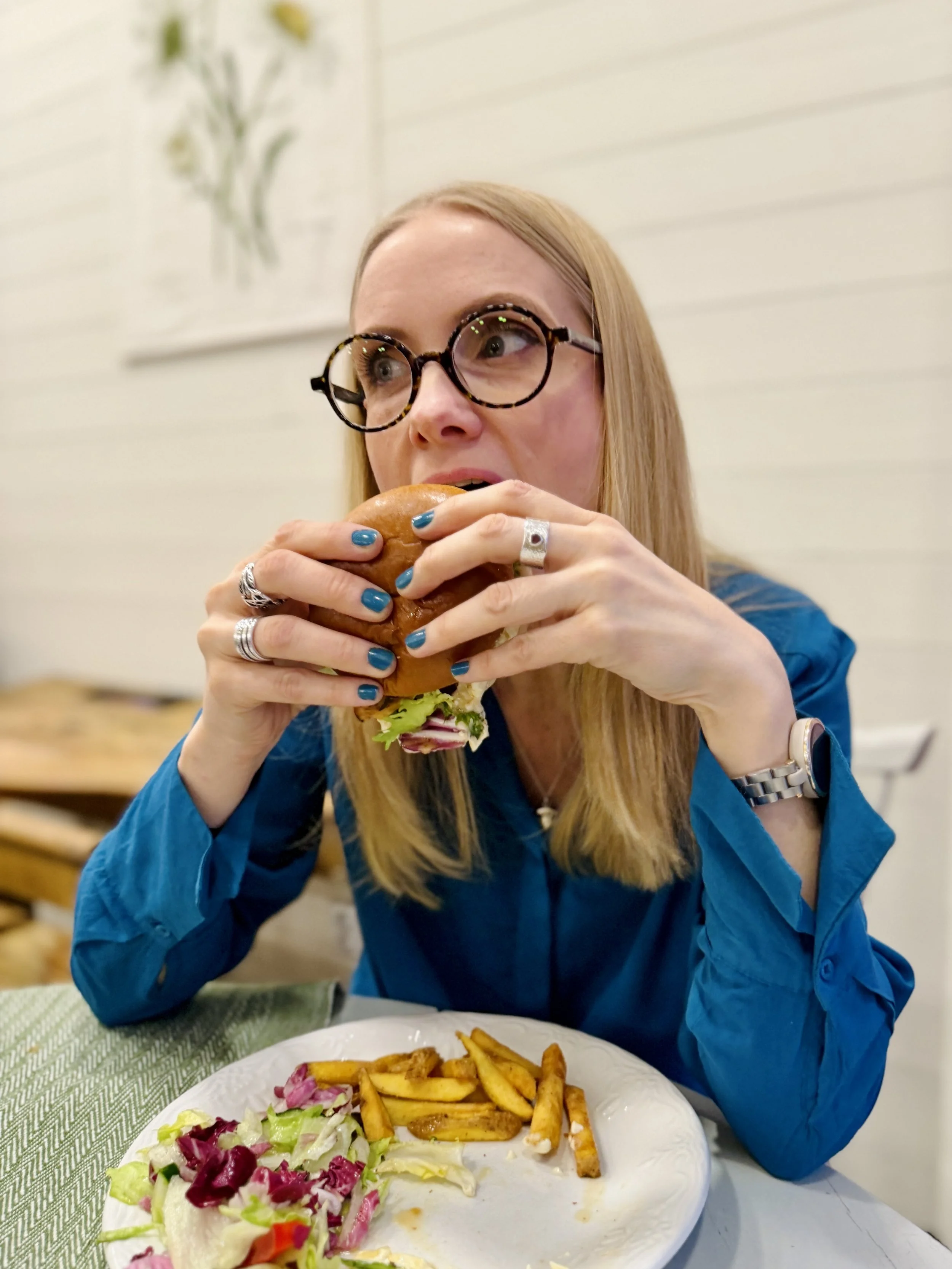 A woman with blonde hair and glasses holding a sandwich close to her mouth while sitting at a table with fries and salad on a white plate.
