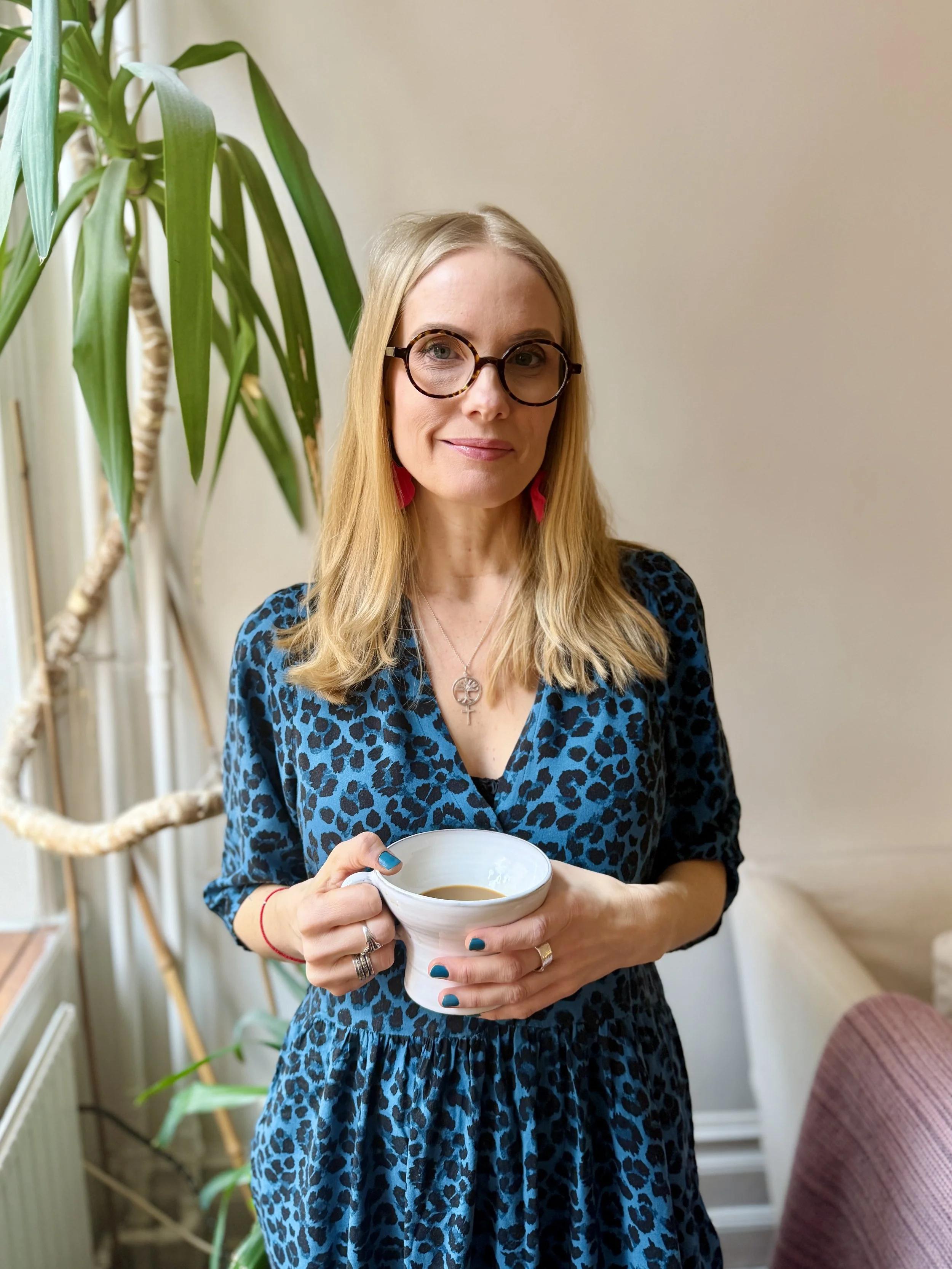 A woman with blonde hair, glasses, and pink earrings standing indoors near a large leafy plant, holding a cup of coffee, wearing a blue leopard-print dress.