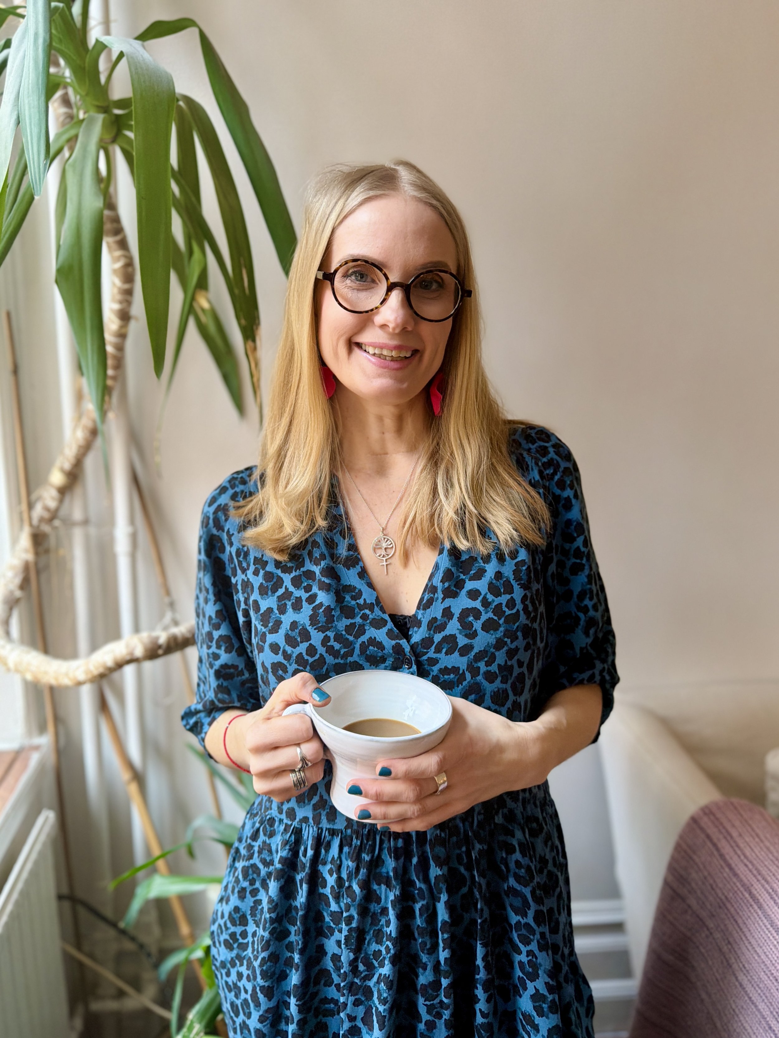 A woman wearing glasses and a blue leopard print dress, holding a cup of coffee, standing indoors near a large green plant.