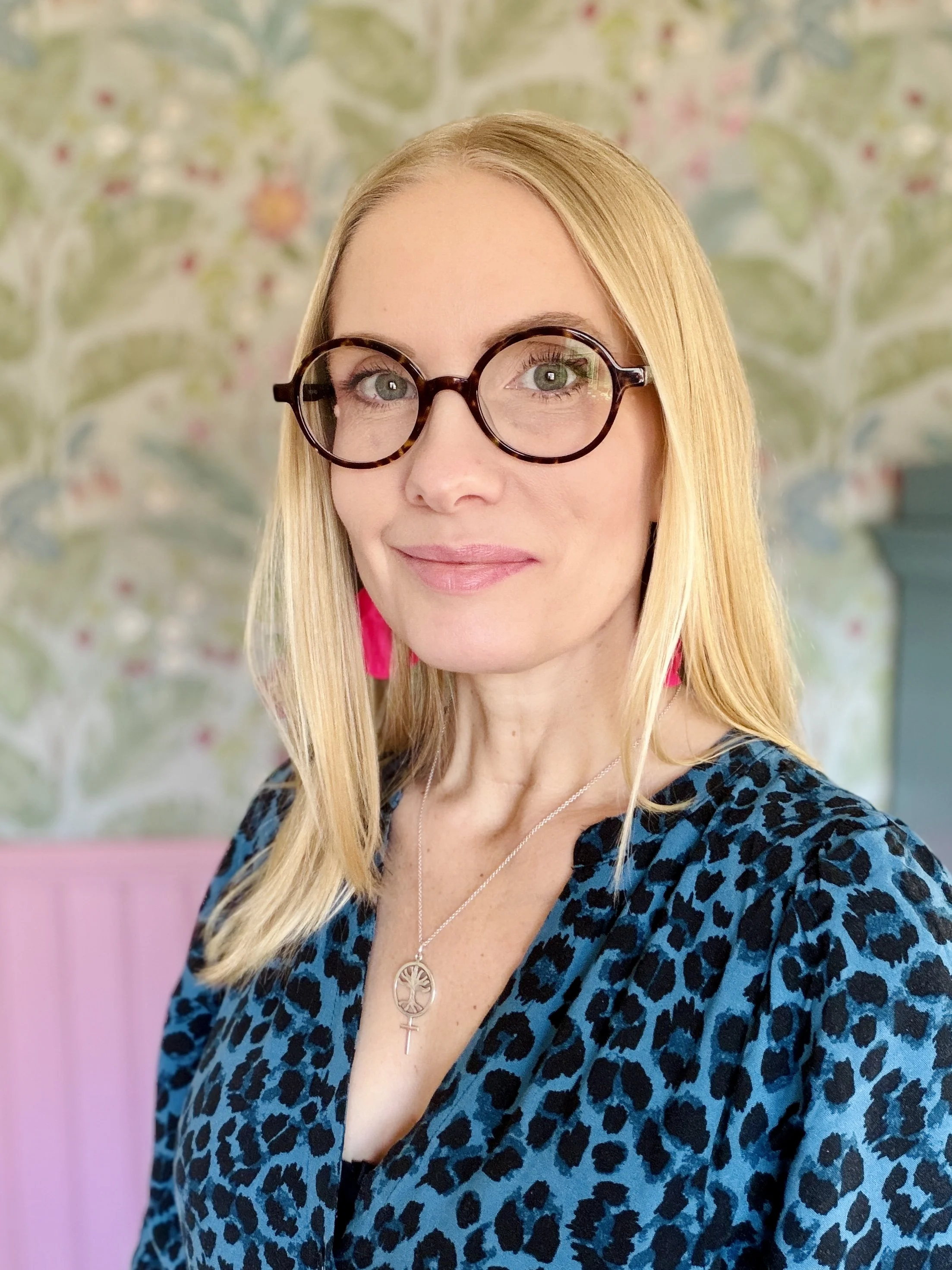 A woman with blonde hair and glasses wearing a blue leopard print top and pink earrings, standing in front of a floral wallpaper background.