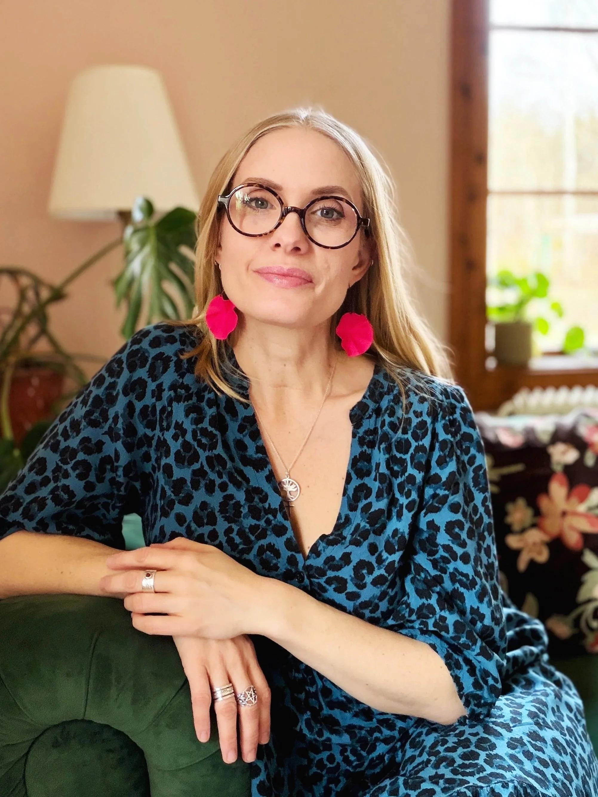 A woman with blonde hair and glasses wearing a blue animal print dress sitting on a green couch, with pink earrings, a necklace, and rings, in a cozy room with a lamp and window with plants.