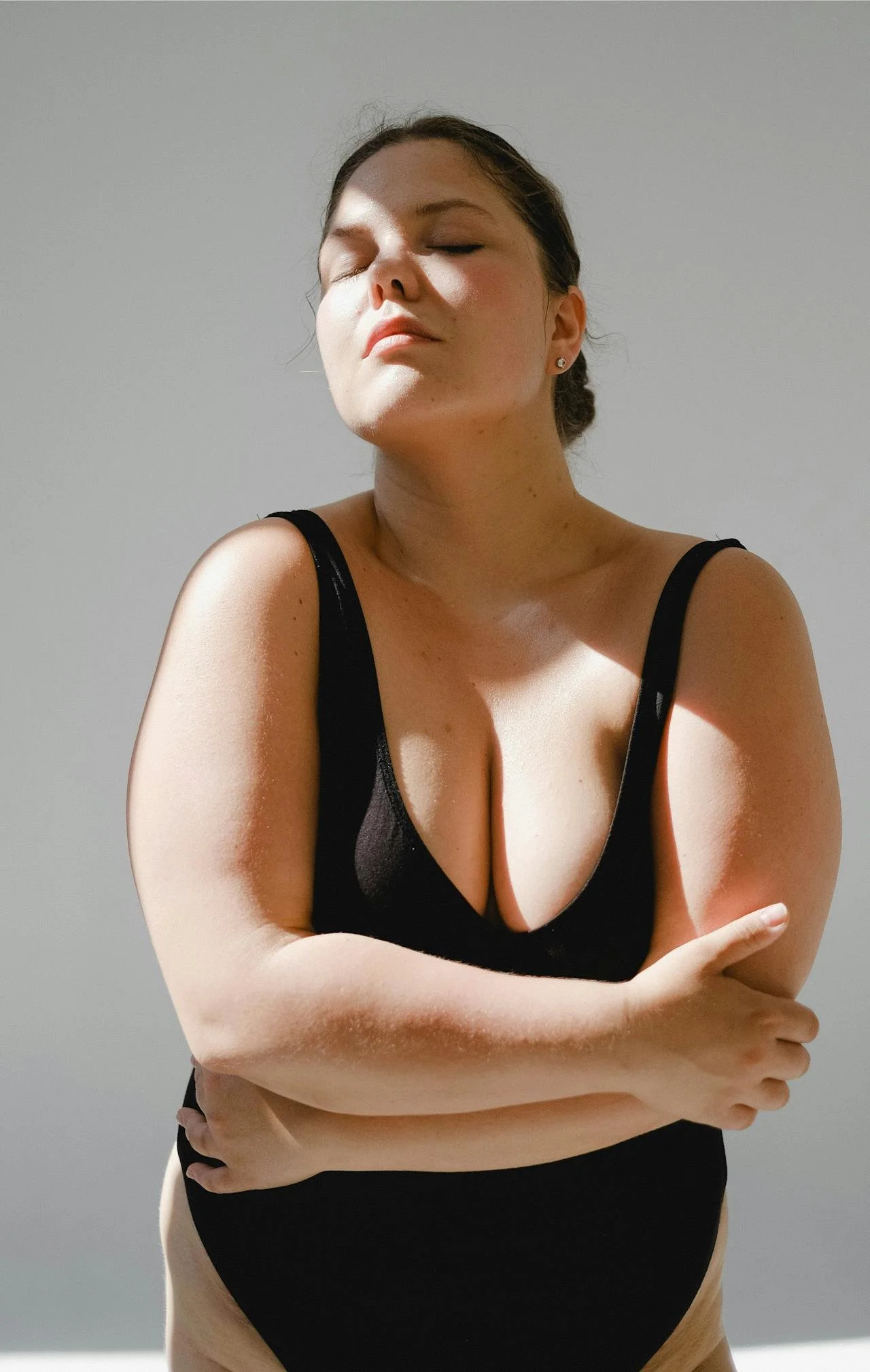 A woman in a black top with thick straps, arm crossed, with her eyes closed and face slightly tilted upwards, standing against a plain sky background.