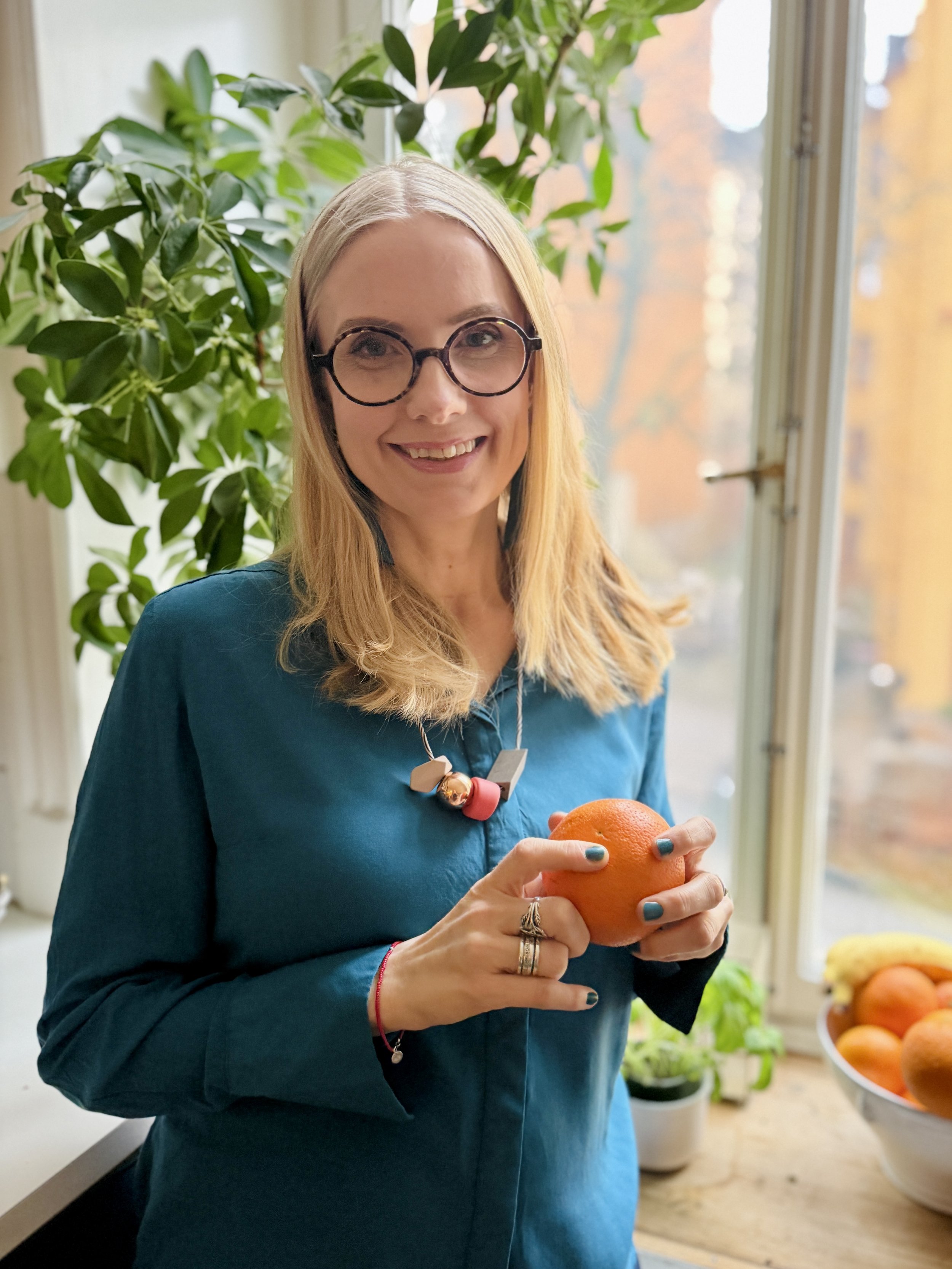 A woman with blonde hair, glasses, and a blue blouse stands by a window holding an orange. There is greenery behind her and a bowl of oranges on the table.