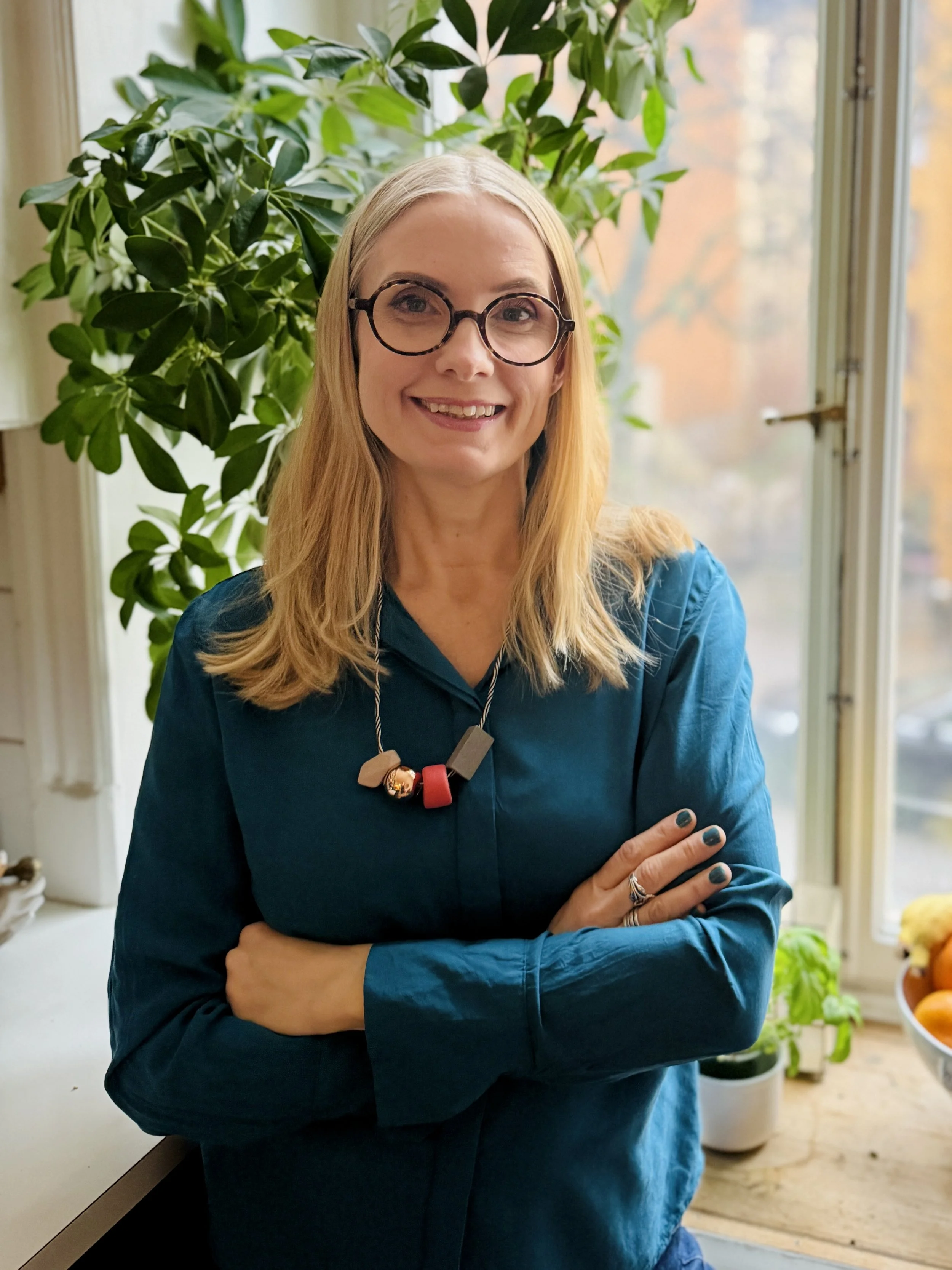 A woman with blonde hair wearing glasses and a blue top, standing indoors near a window with a large green plant behind her. She is smiling and has her arms crossed.