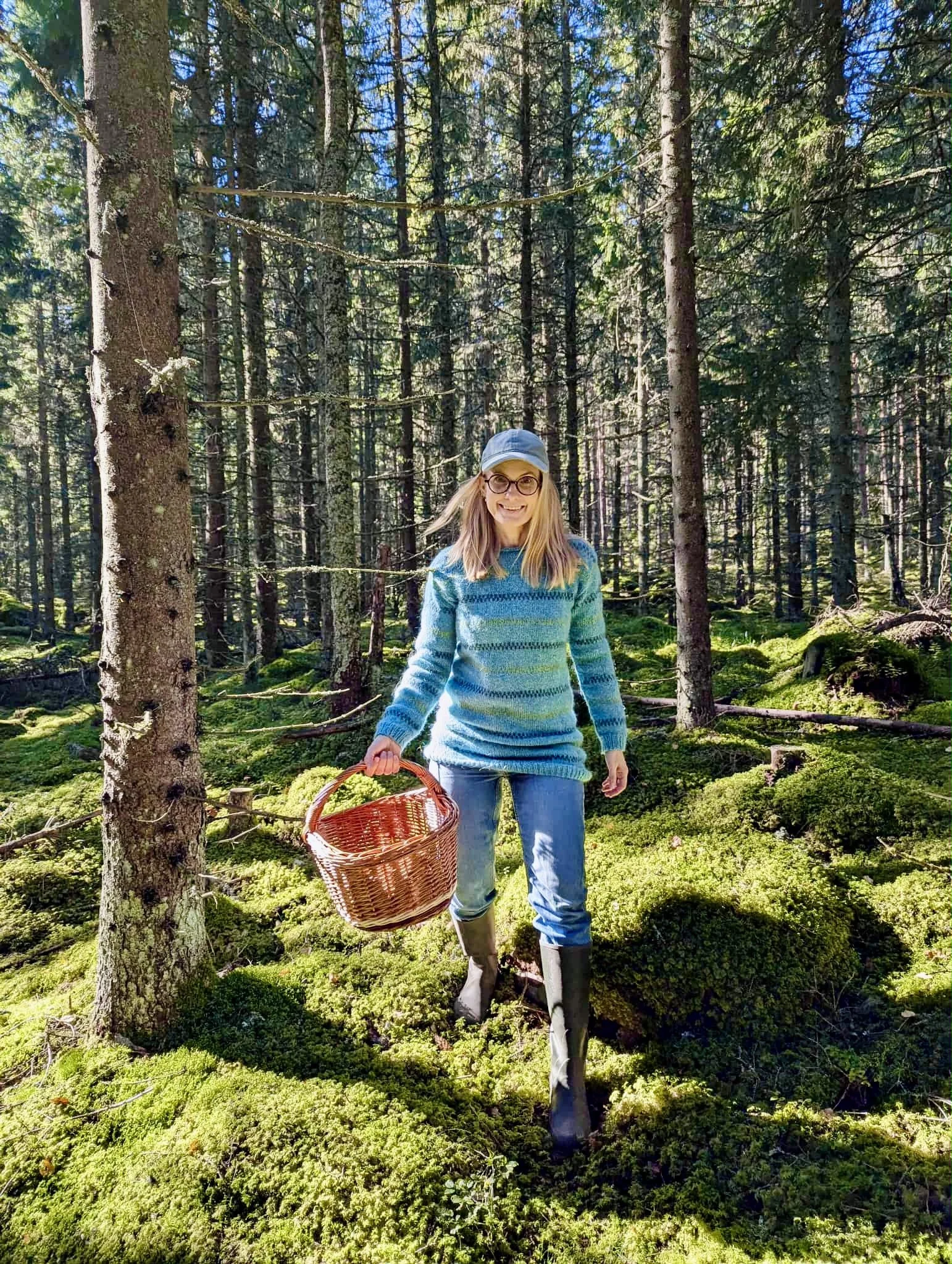 A woman with glasses, a light blue cap, a blue striped sweater, and jeans, carrying a wicker basket, walking through a dense forest with tall trees and green moss-covered ground.