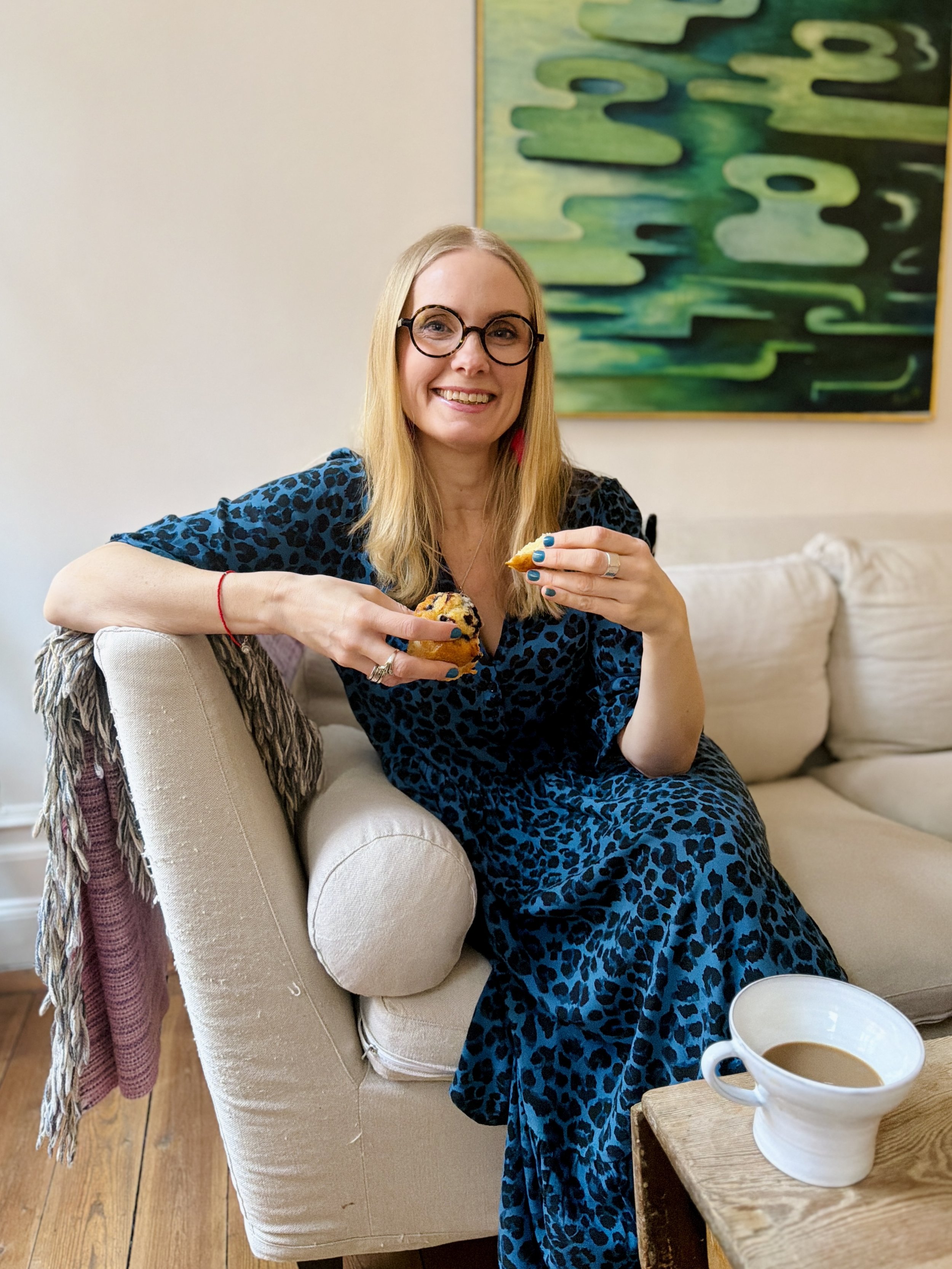 A woman with blonde hair, glasses, and a blue animal print dress sitting on a beige sofa, holding a muffin and a spoon, smiling at the camera. There is a cup of coffee on a wooden table in front of her, and a colorful abstract painting hangs on the wall behind her.