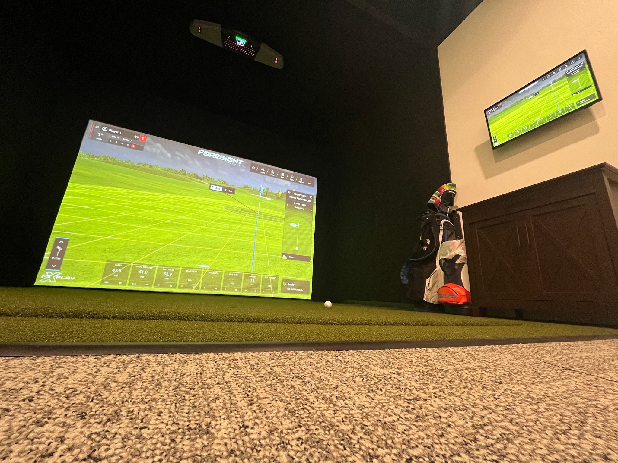 Indoor golf simulator room with a large screen displaying a virtual golf course, a smaller screen on the wall showing the same course, a golf club headcover, a golf bag, and a golf ball on artificial turf.