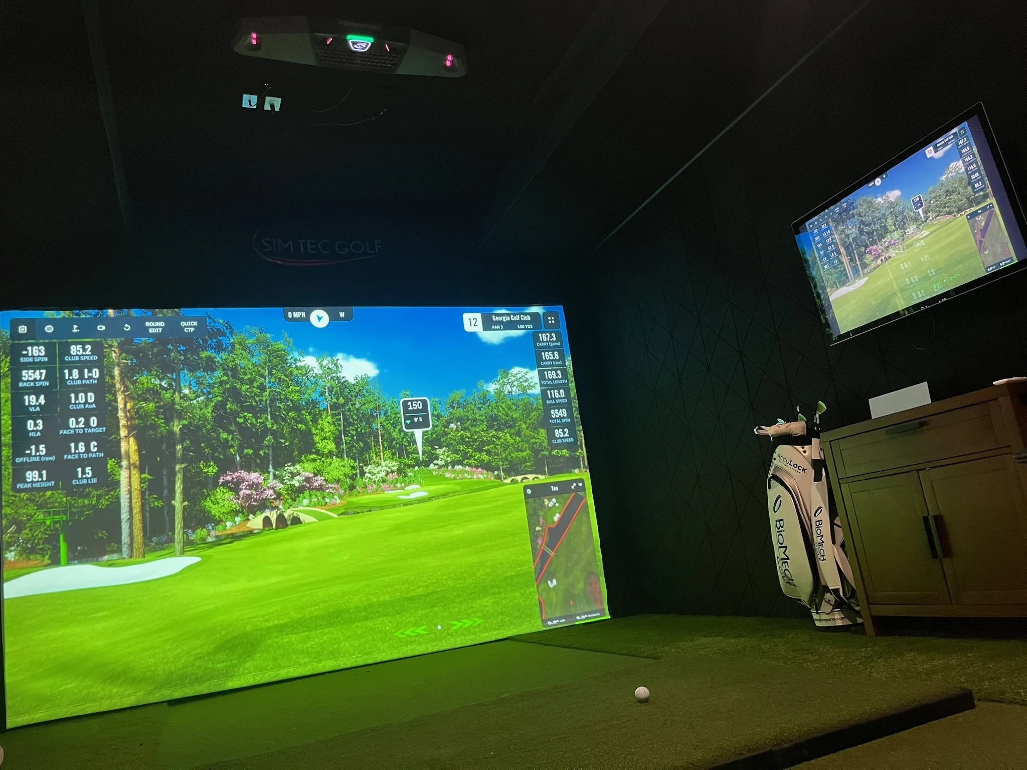 Indoor golf simulator room with a large screen displaying a virtual golf course, a smaller screen showing the course map, golf clubs in a bag, and a golf ball on a mat.