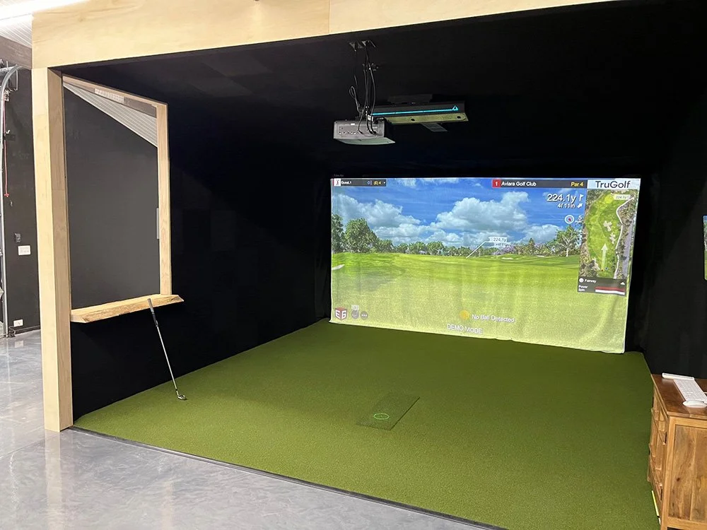 Indoor golf simulation room with a large screen displaying a golf course, a green mat for swinging, a golf club leaning against a wooden enclosure on the left, and a small wooden side table on the right.