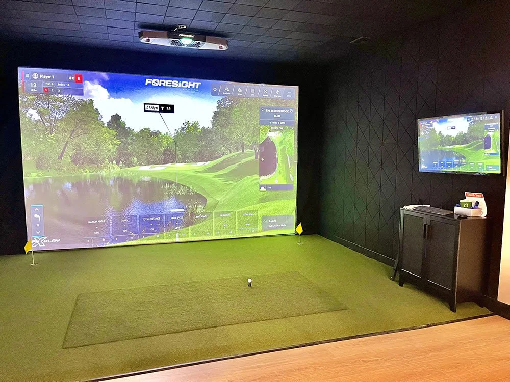 Indoor golf simulator room with a large screen displaying a golf course, a small putting green with a golf ball and two flags, and a smaller TV screen on the right.