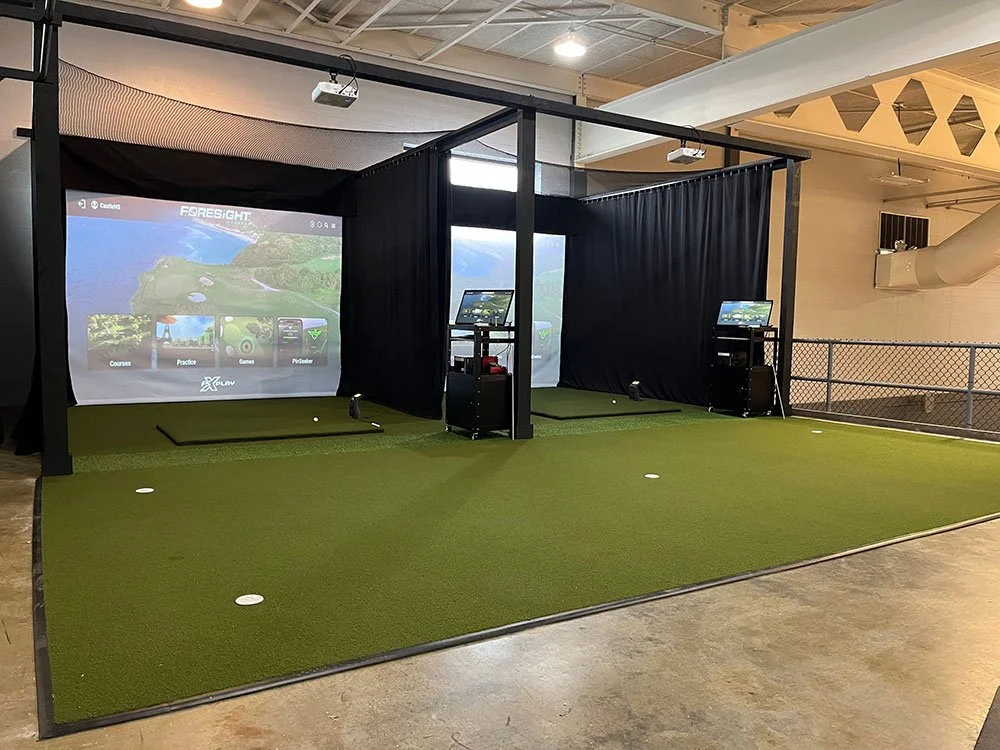 Indoor golf simulator setup with two hitting bays, artificial turf, and large screens displaying virtual golf courses.