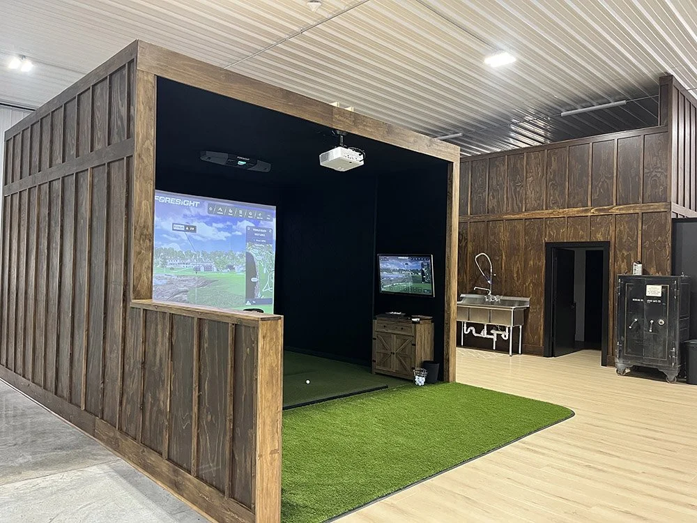 Indoor golf simulator setup with a large screen, small television, and golf mat, enclosed in wooden walls, with a sink and storage area nearby.