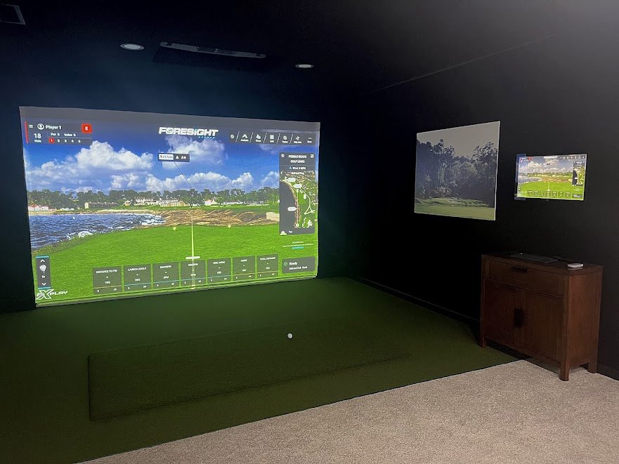Indoor golf simulation room with a large screen displaying a golf course, two smaller monitors showing golf scenery, a virtual golf ball on the green, and a wooden cabinet in the corner.