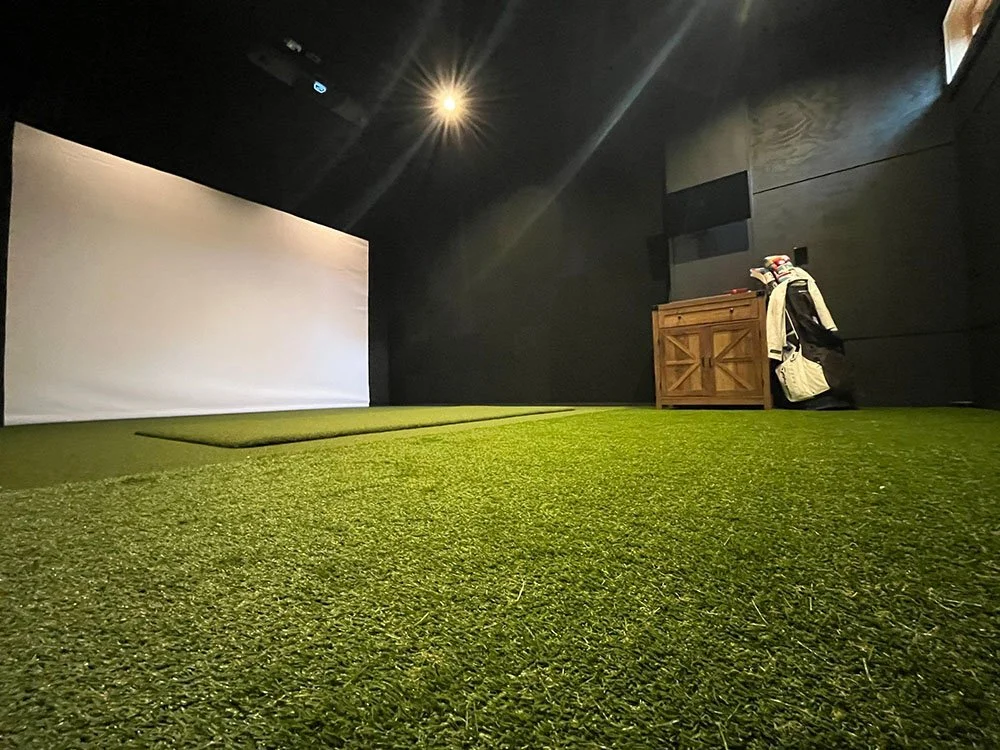 Indoor golf simulator room with artificial turf, a large white screen, a wooden storage unit, golf bags, and club sets against black walls.