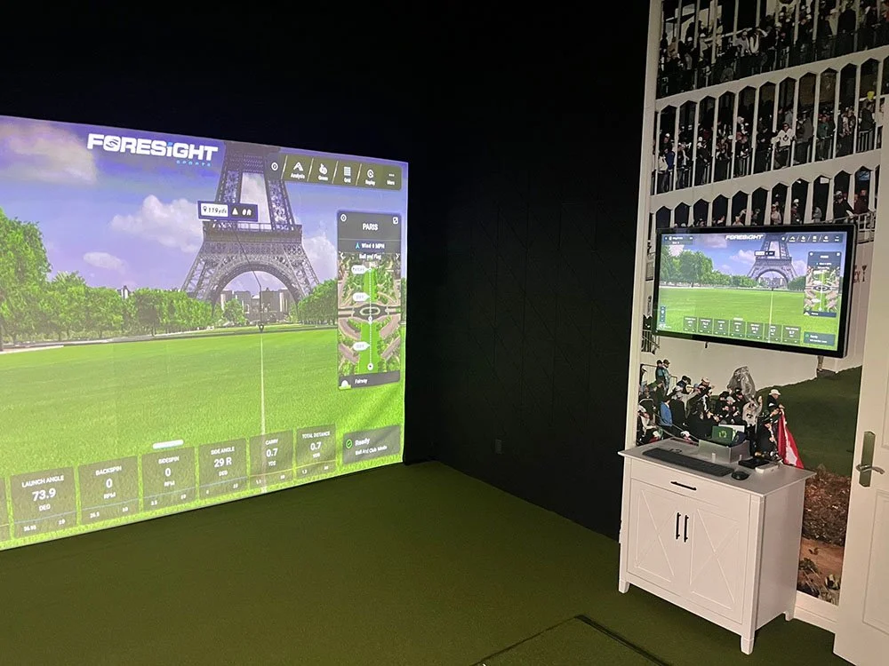 Indoor golf simulation room with digital screens displaying a golf course, one large screen on the left and a smaller one on the right, featuring the Eiffel Tower background and golf data.