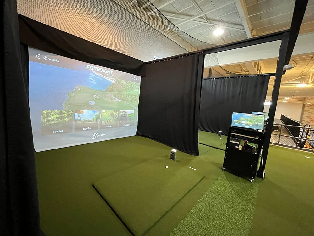 Indoor golf simulator setup with a large screen displaying a golf course, a golf mat, a golf ball, a PC with a monitor, and black curtains for privacy.