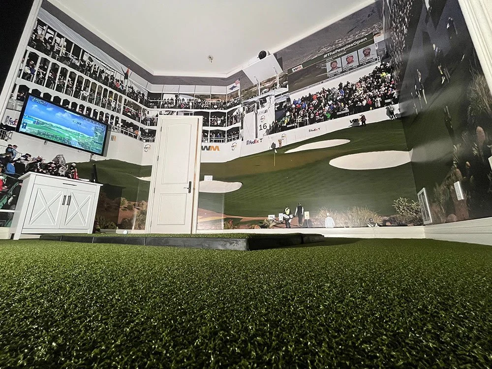 A room with a golf-themed mural on the wall, depicting a golf course and spectators, a television showing a golf game, a white cabinet, and a door. The floor has green artificial turf.