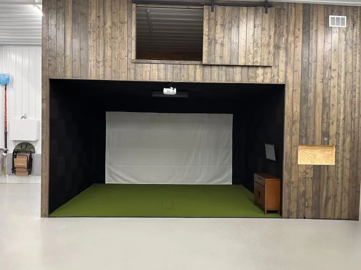 Indoor golf simulation room with a green artificial turf floor, a white screen at the back, and a projector mounted on the ceiling.