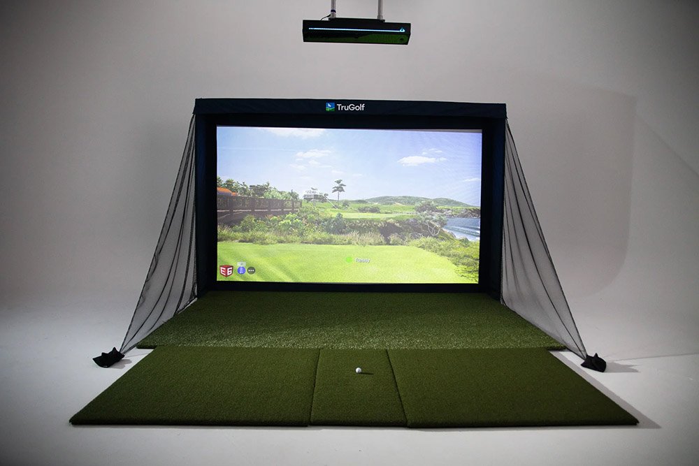 Indoor golf simulator setup with a large screen displaying a golf course, surrounded by black curtains, with green turf mats and a golf ball in front.