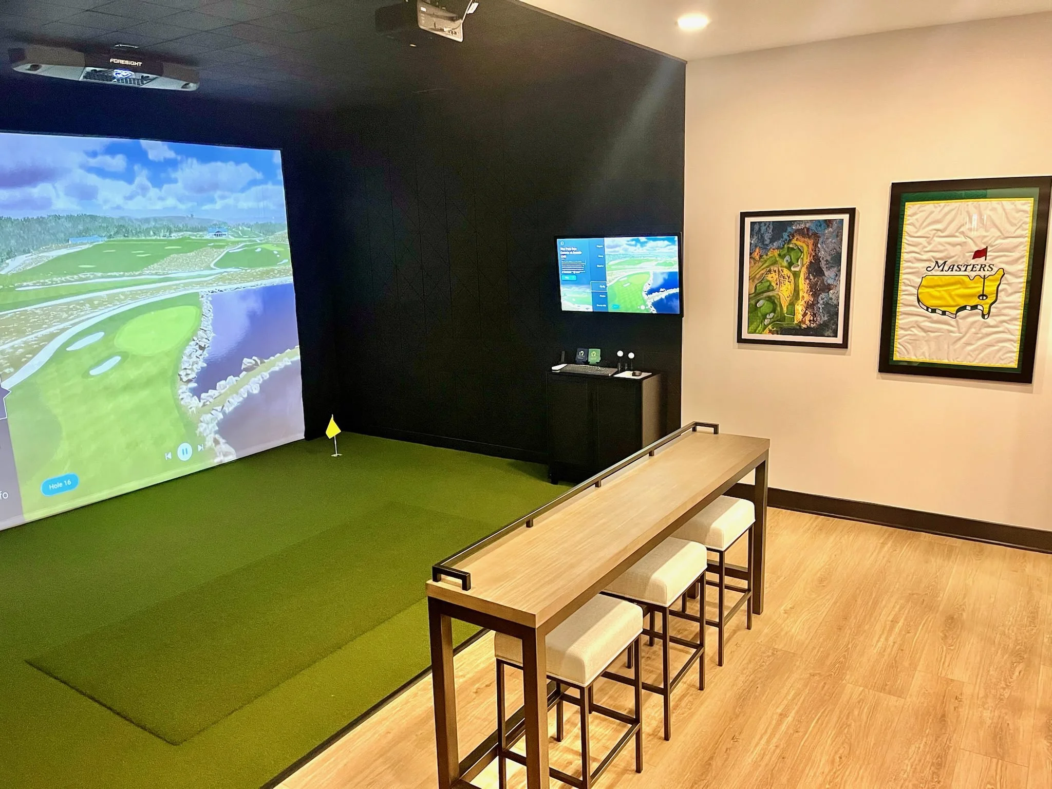 Indoor golf simulation room with large digital screen displaying a golf course, high-top table with four chairs, two smaller screens on the wall, and framed golf-themed artwork.