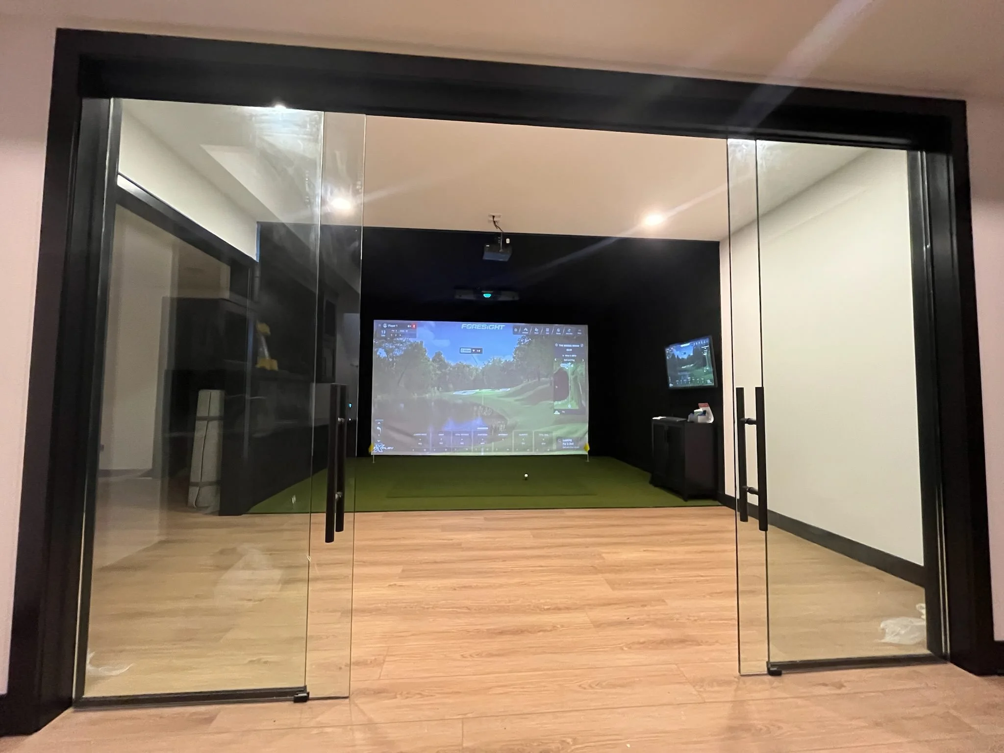 Indoor golf simulator room with glass sliding doors, a large screen displaying a golf course, and a smaller screen on the right, with a green turf area and wooden flooring.