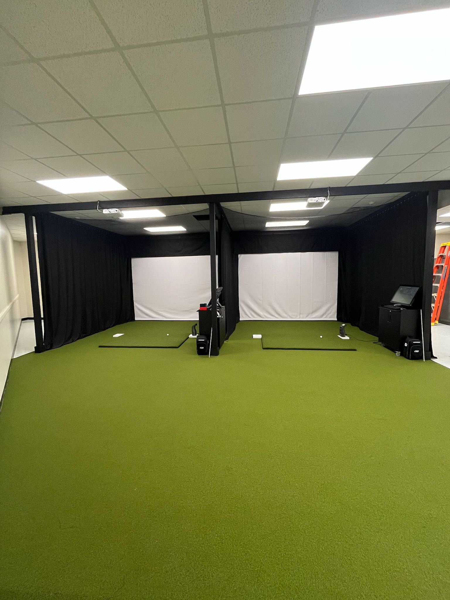 Indoor golf simulator room with green artificial turf, black curtains, white screens, and golf equipment.
