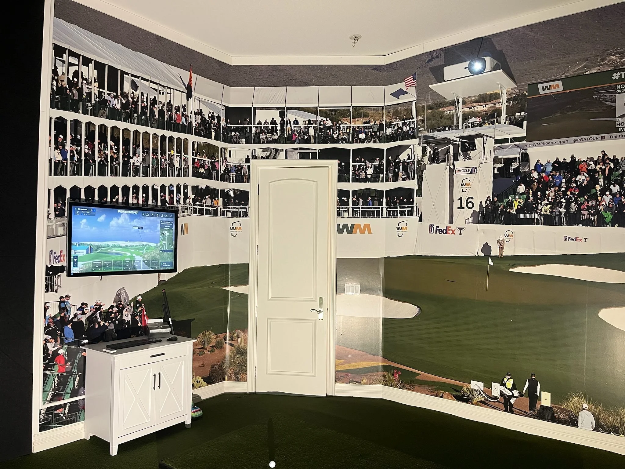 Indoor golf simulation room with a large wall mural of a golf course, a television screen displaying a golf game, and a door in front of the mural.